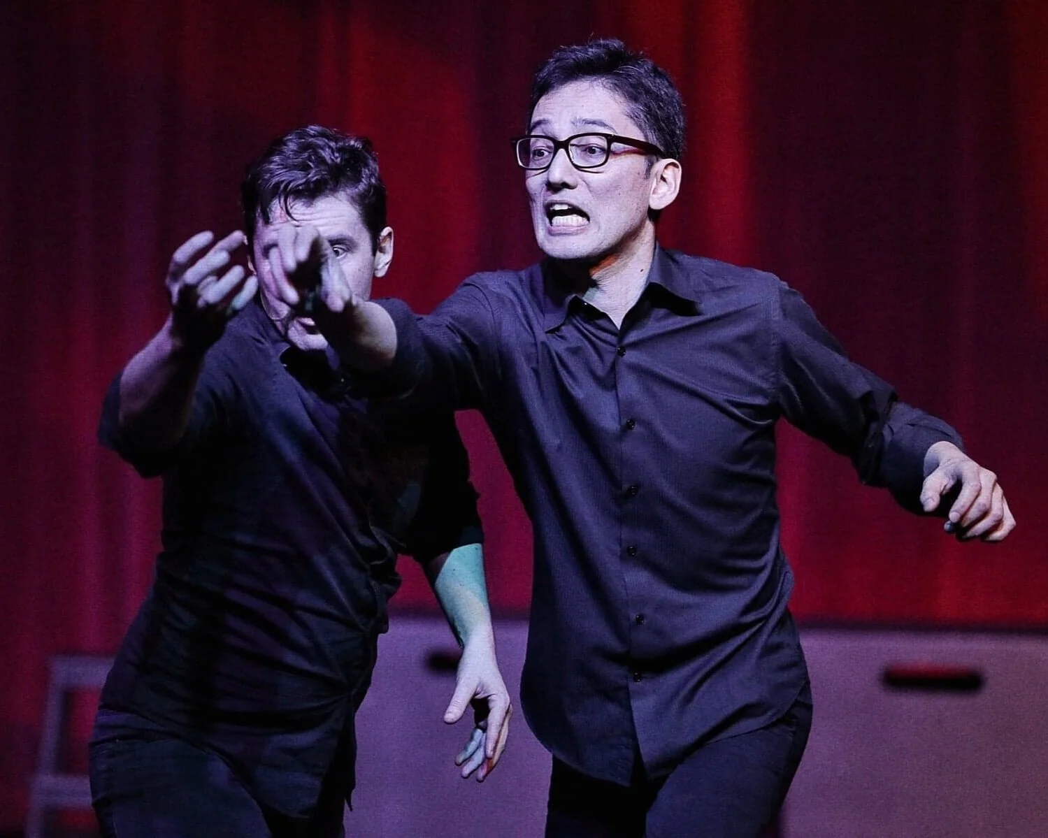 Travis improvising with Chris Ross, both dressed in black, red curtain behind. Travis is blocking Chris's face, and his mouth is open in an exaggerated grimace.