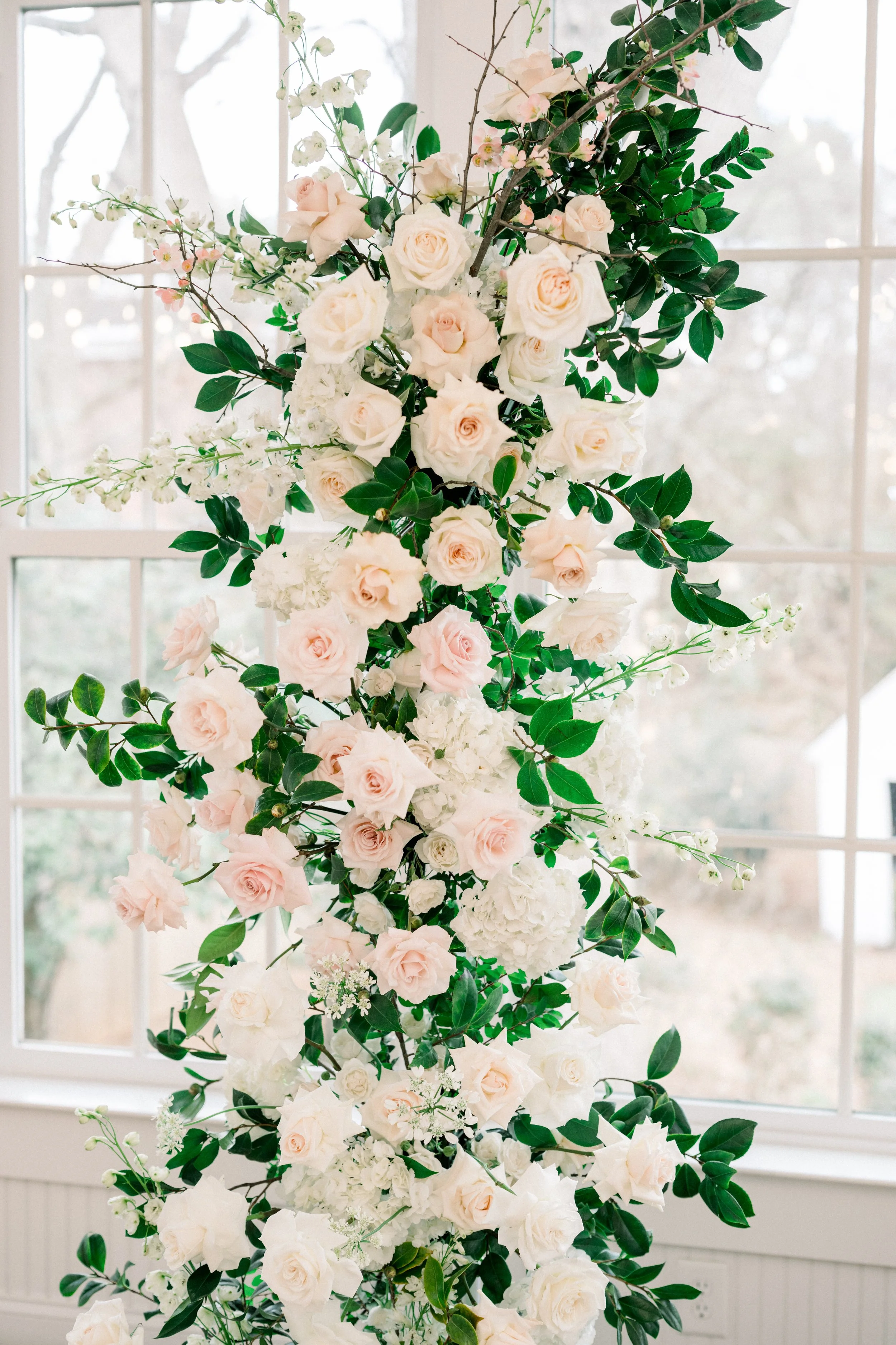 A large floral arrangement with white and pale pink roses, white hydrangeas, and greenery, placed in front of a window with a view of trees outside.