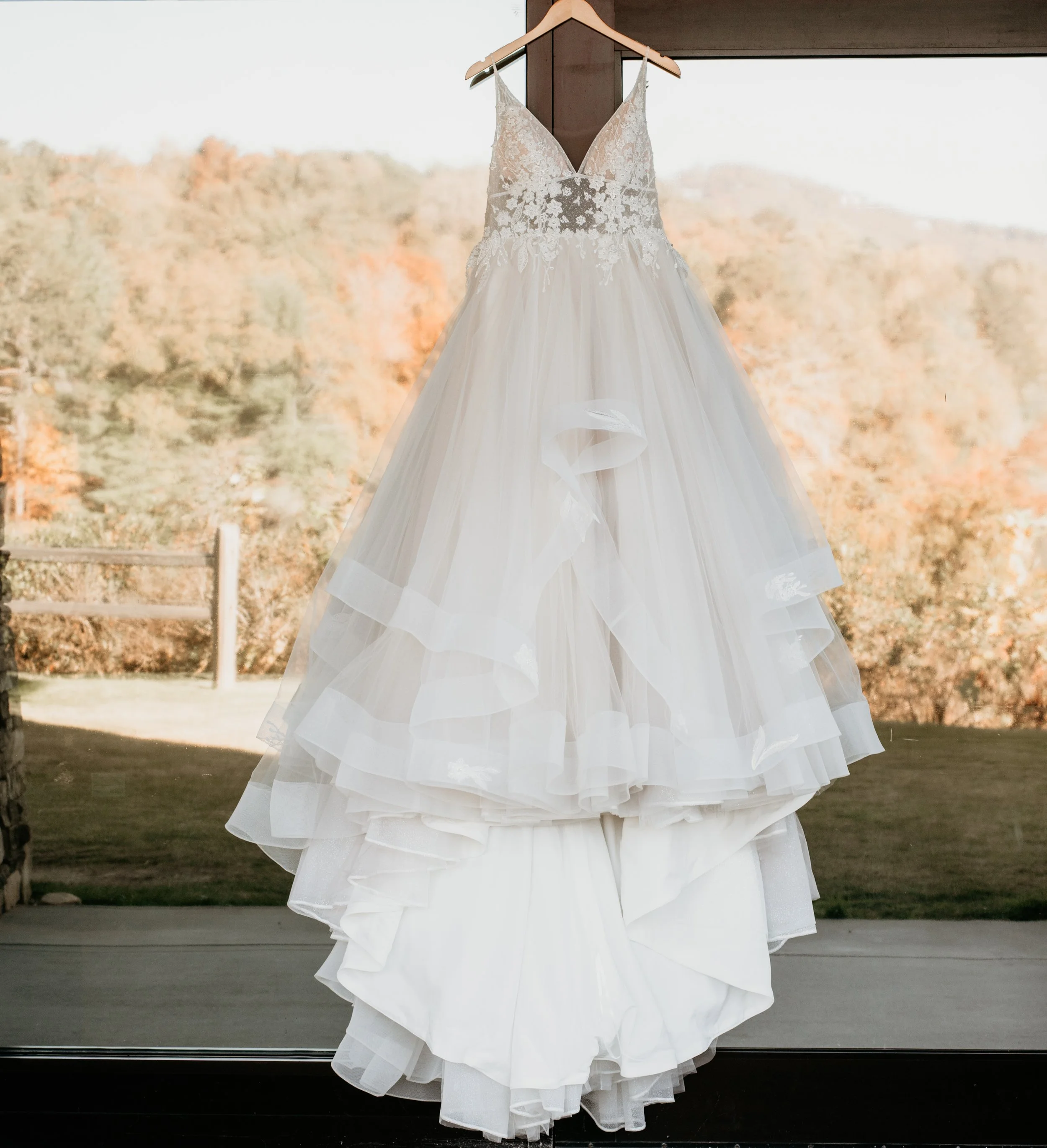 A white wedding dress with lace detailing on the bodice and a layered tulle skirt hanging on a wooden hanger in front of a large window showing a scenic view of trees and mountains in autumn colors.