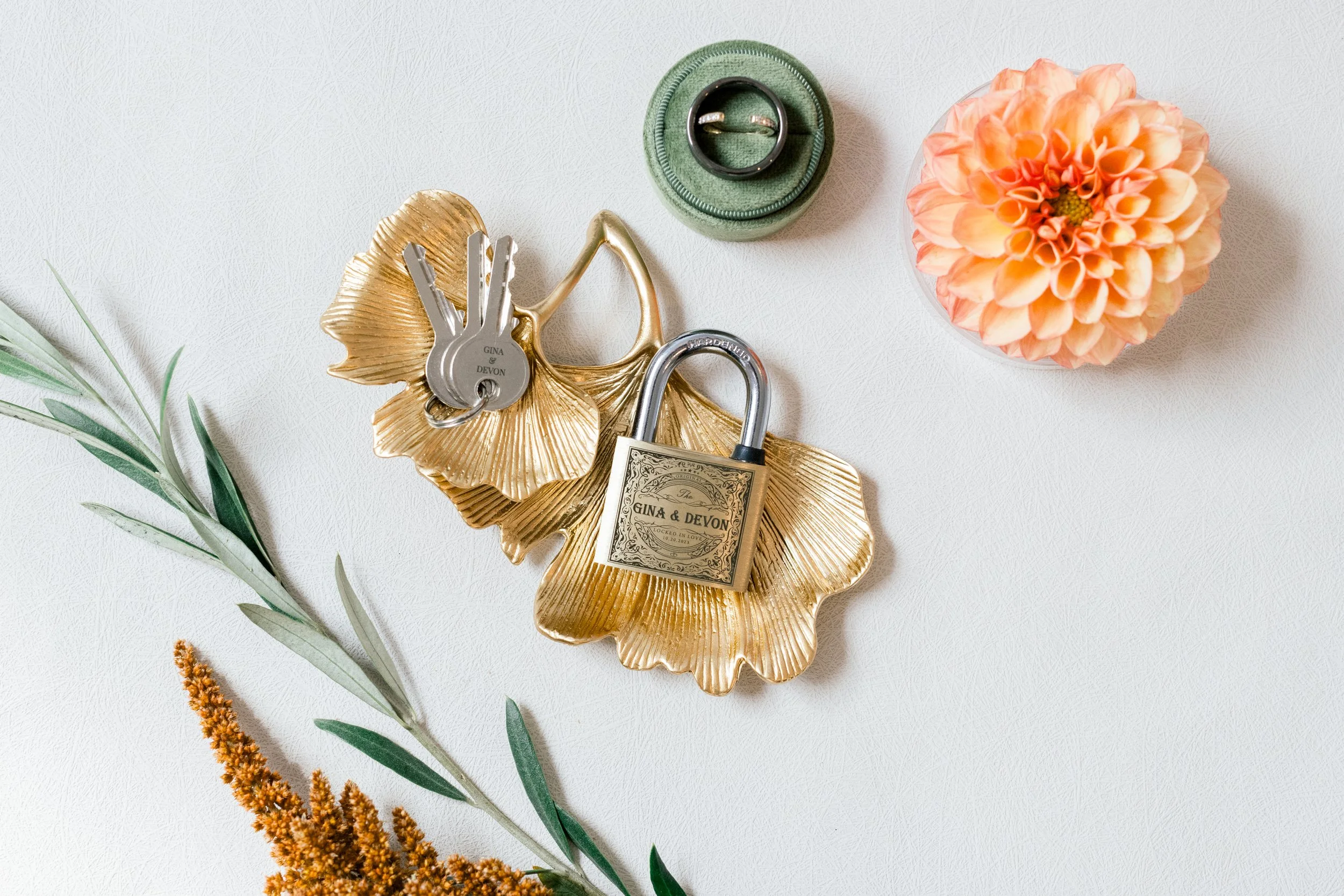 A gold-colored leaf-shaped tray with rings and a lock on it, a closed green ring box, a peach-colored dahlia flower in a glass container, and some green leafy sprigs with orange and brown flowers on a white textured surface.
