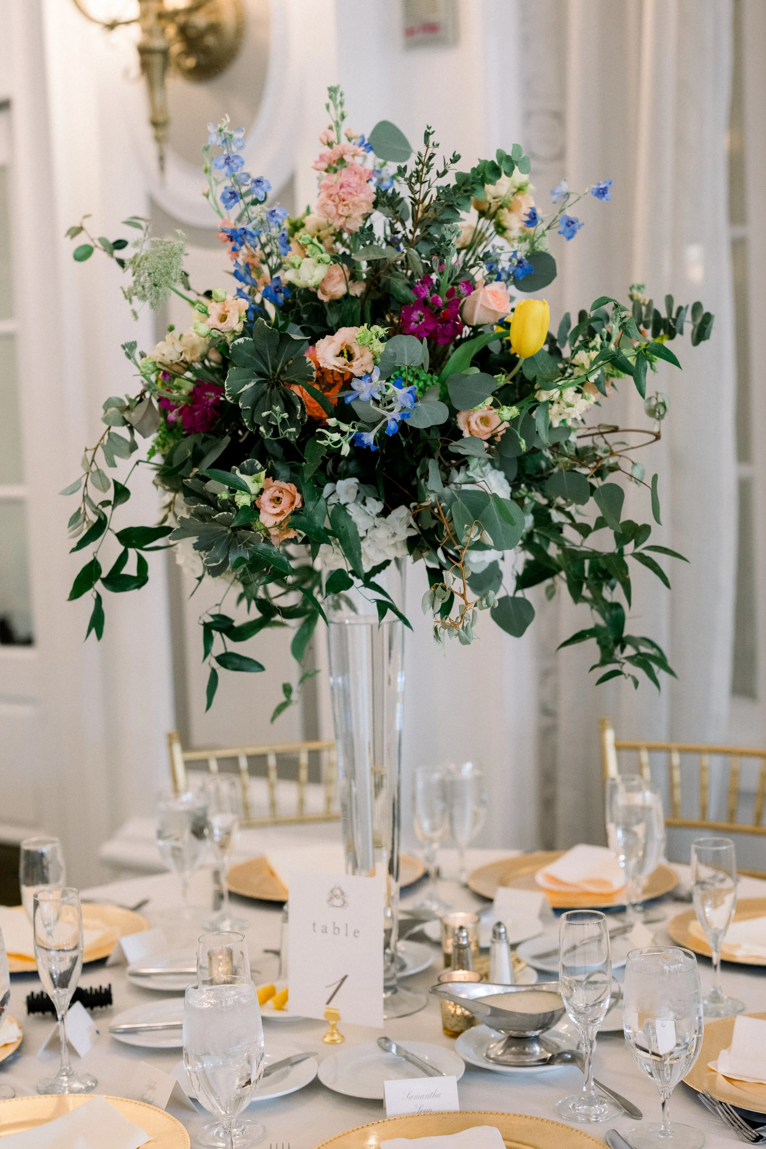 A formal table setting with a tall floral centerpiece featuring pink, blue, yellow, and white flowers and green foliage, surrounded by glassware, plates, silverware, and gold accents in an elegant event space.