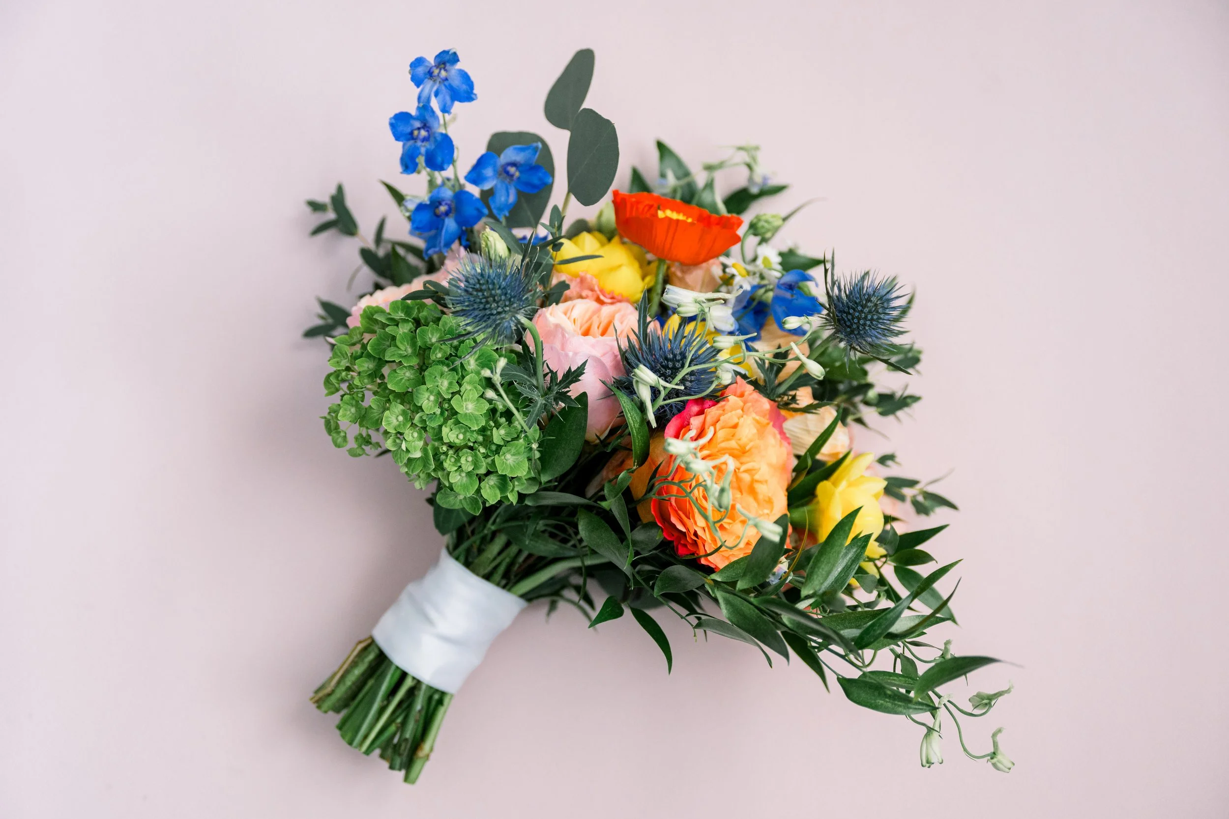 A colorful bouquet of flowers including blue delphiniums, orange poppies, pink roses, yellow flowers, and green foliage, wrapped with white ribbon.