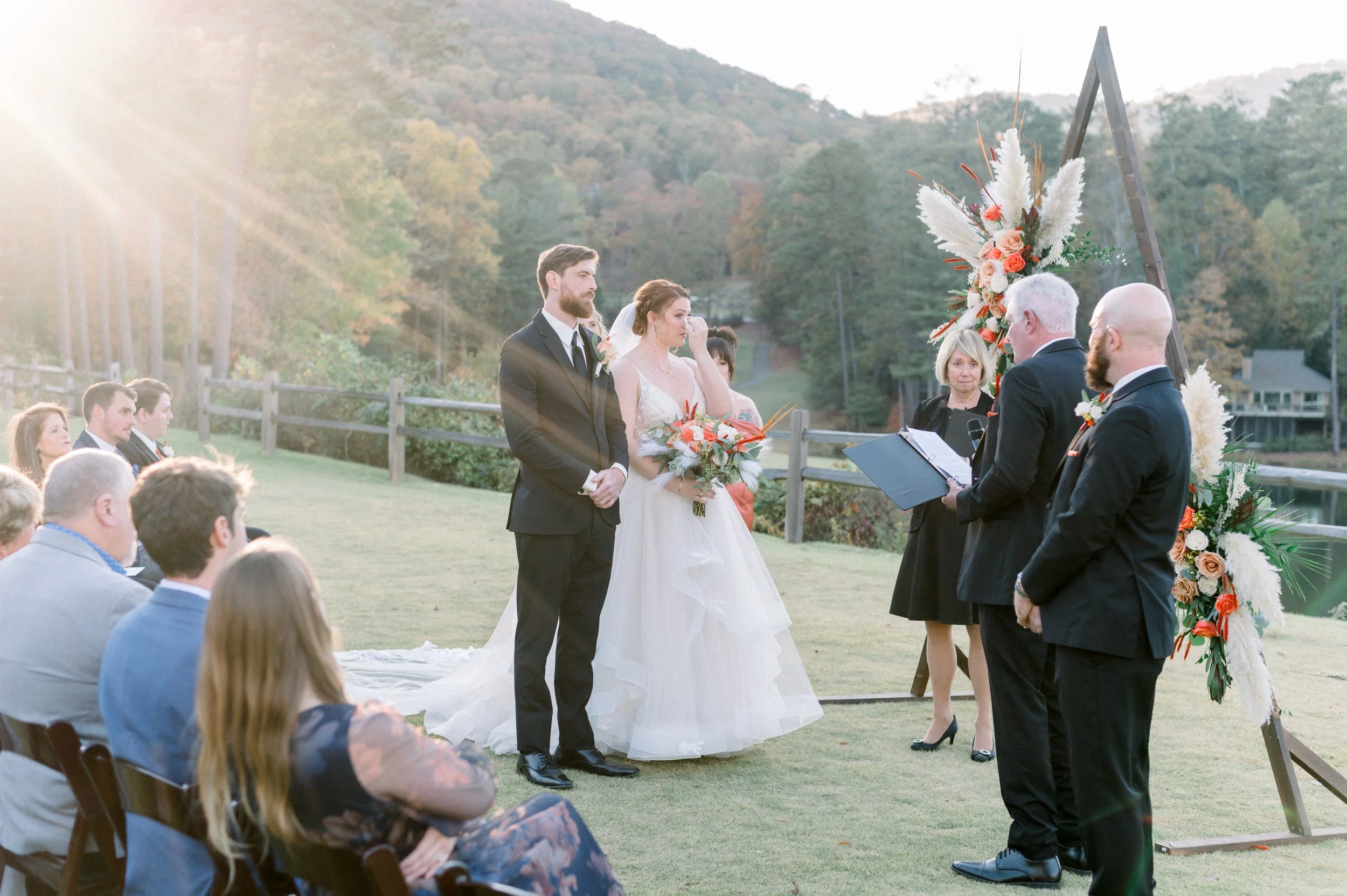 A couple getting married outdoors with the officiant and guests watching. The groom is wearing a black suit, and the bride is in a white wedding dress holding a bouquet. There is a floral arrangement with white and peach-colored flowers on a wooden t