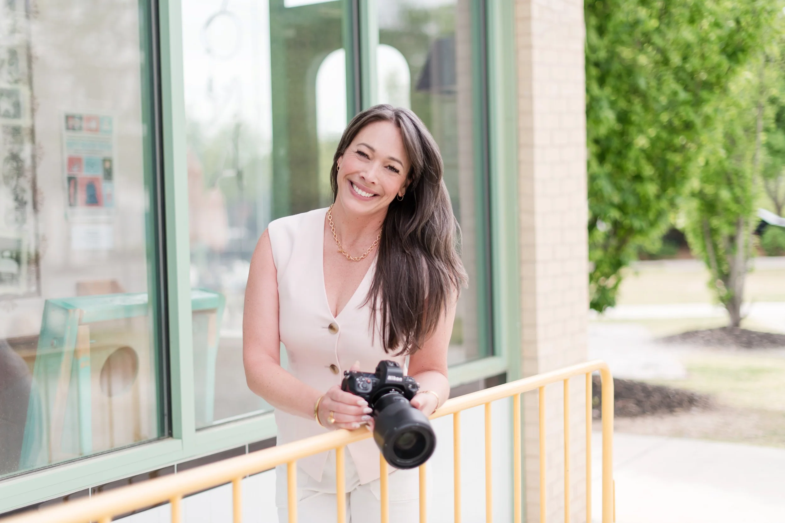 A smiling woman with long brown hair wearing a light-colored sleeveless dress and holding a camera, standing outside near a glass window with green trees in the background.