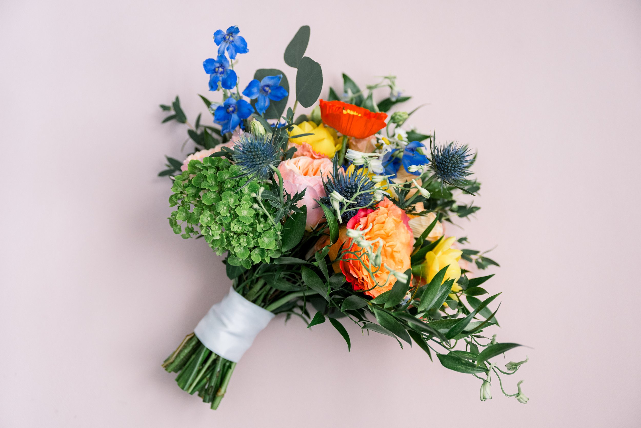 Bouquet of mixed flowers including blue delphiniums, orange poppies, pink roses, yellow flowers, and various greenery tied with white ribbon.