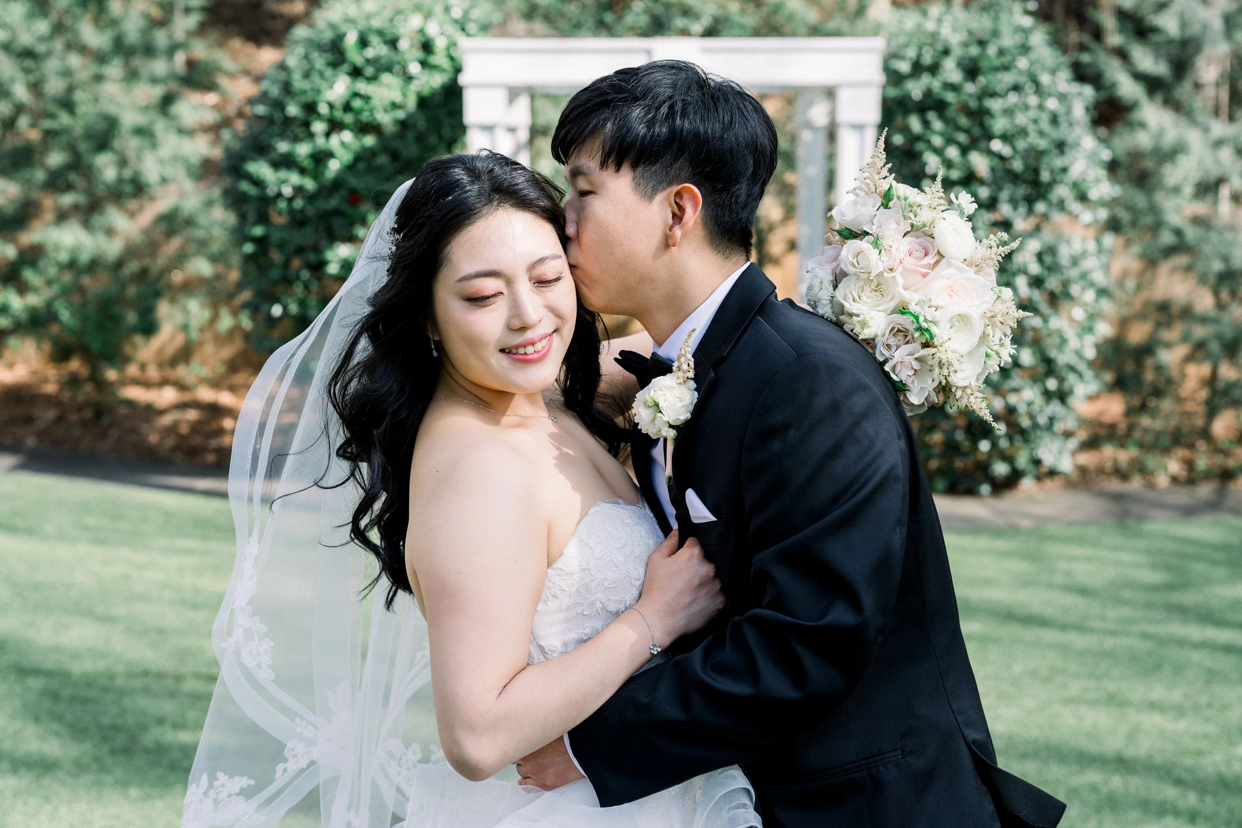 A bride and groom share a kiss outdoors, with the groom holding a bouquet of white and pink flowers behind him. The bride is wearing a white wedding dress and veil, the groom is wearing a black tuxedo, and there is greenery in the background.
