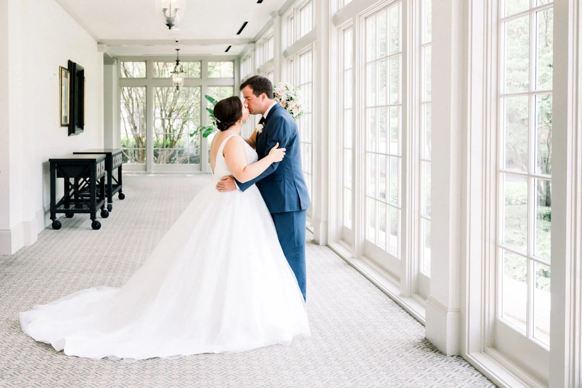 A bride and groom sharing a kiss inside a bright, airy wedding venue with large windows and white walls.