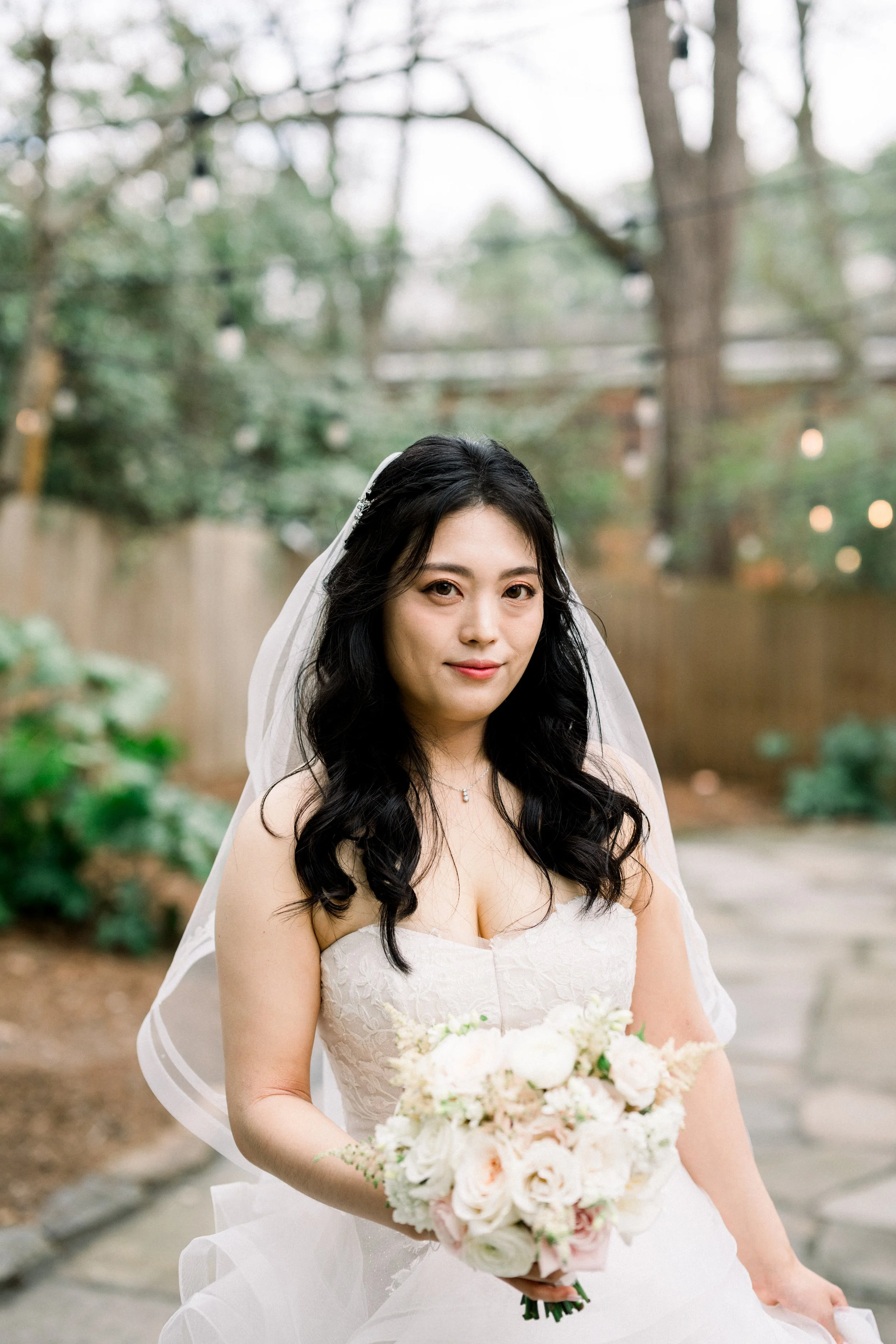 A bride holding a bouquet of white and pink flowers, standing outdoors with trees and string lights in the background.