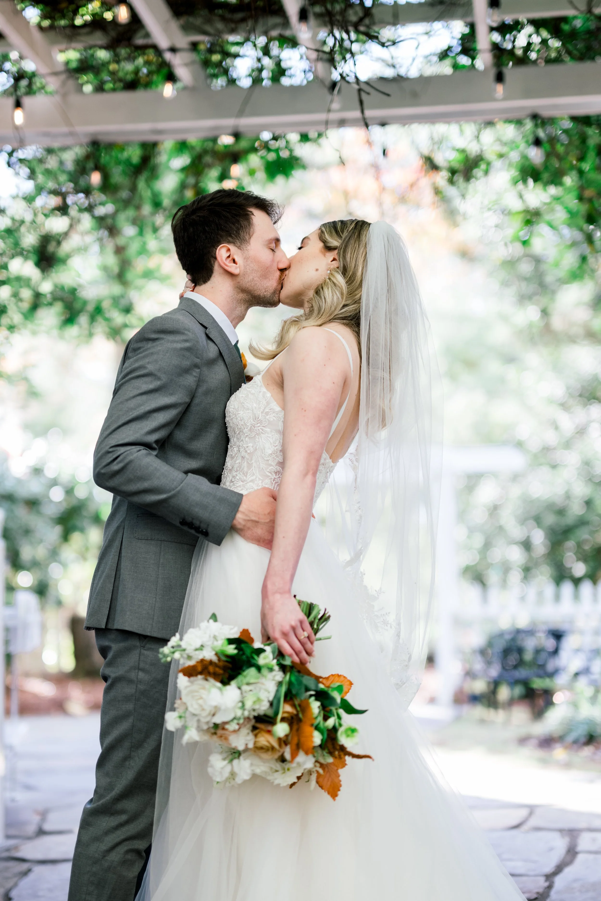 A bride and groom sharing a kiss during their outdoor wedding ceremony. The bride is wearing a white wedding dress and veil, holding a bouquet of flowers. The groom is dressed in a gray suit. Trees and string lights are in the background.