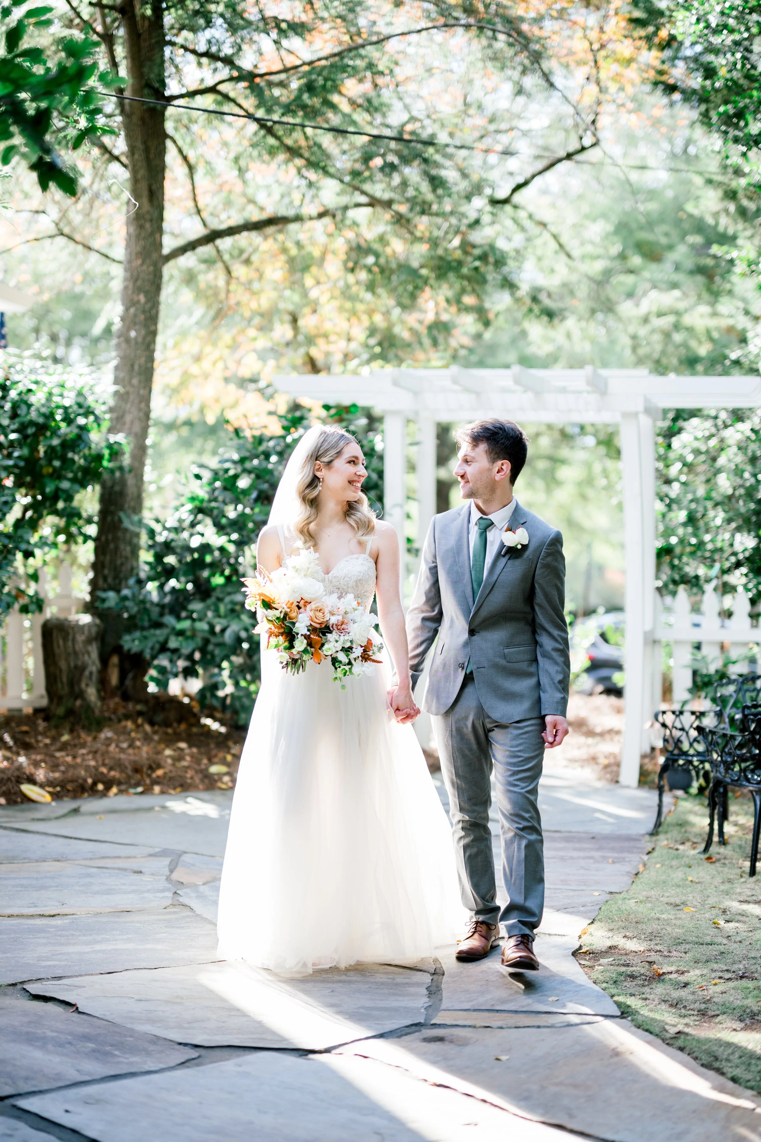 A bride and groom walking hand in hand outdoors during their wedding ceremony, smiling at each other surrounded by trees and greenery.