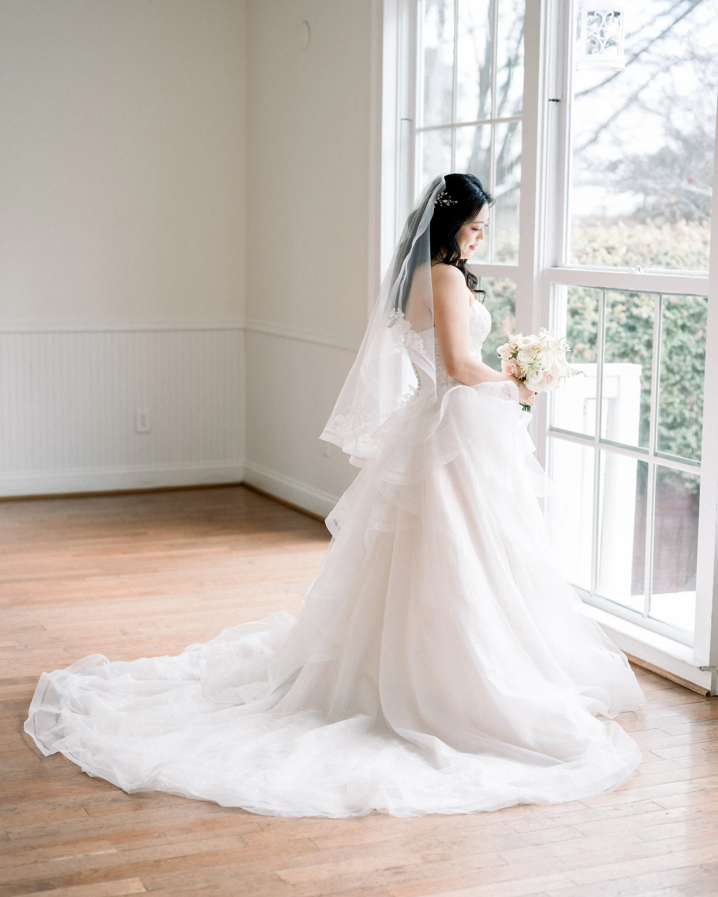 A bride in a white wedding dress standing near a large window, holding a bouquet, with her veil flowing onto the wooden floor.