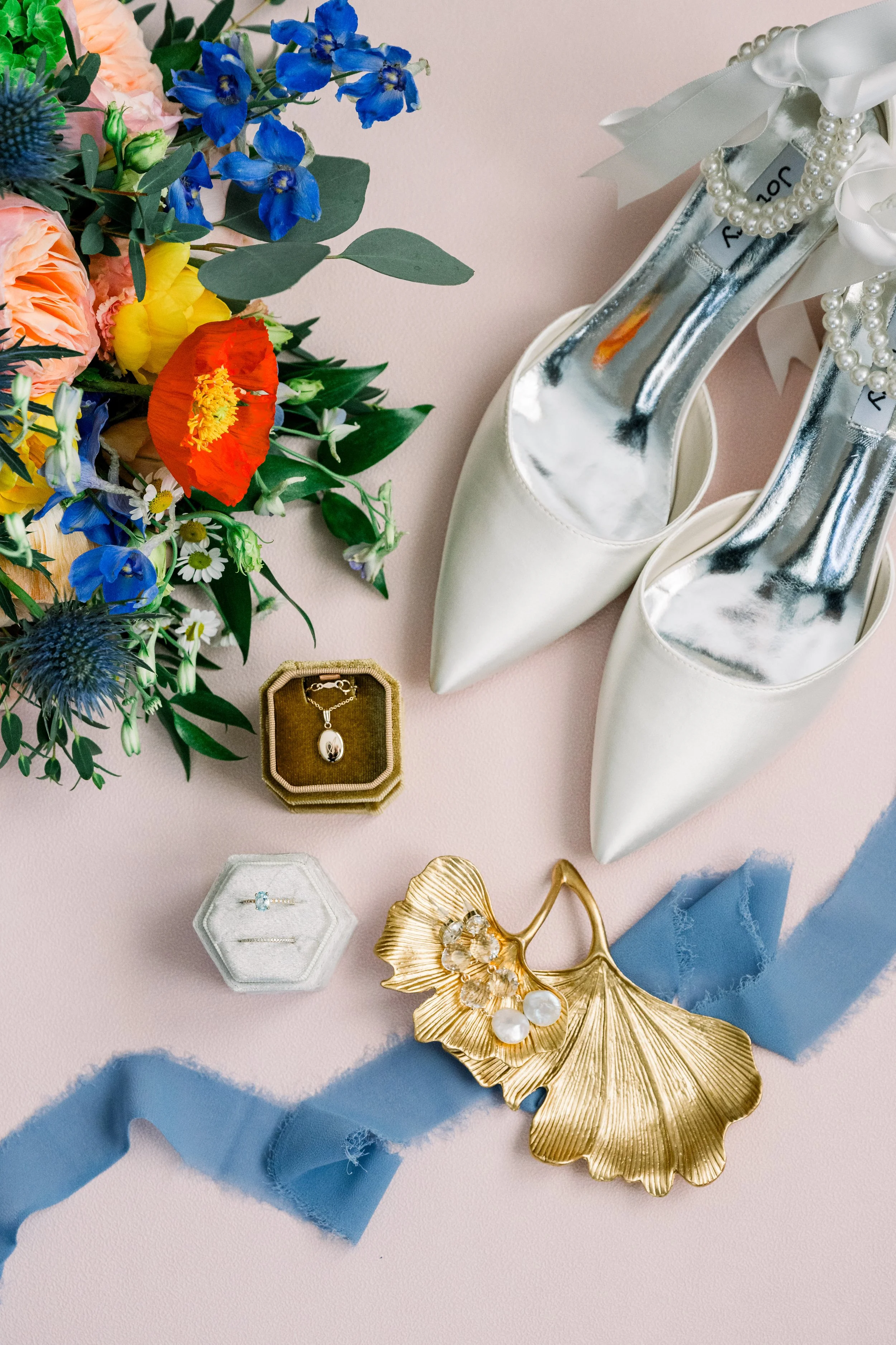 A bouquet of colorful flowers, white high heels with pearl accents, and jewelry including a necklace and rings on a pink surface with blue tissue paper.