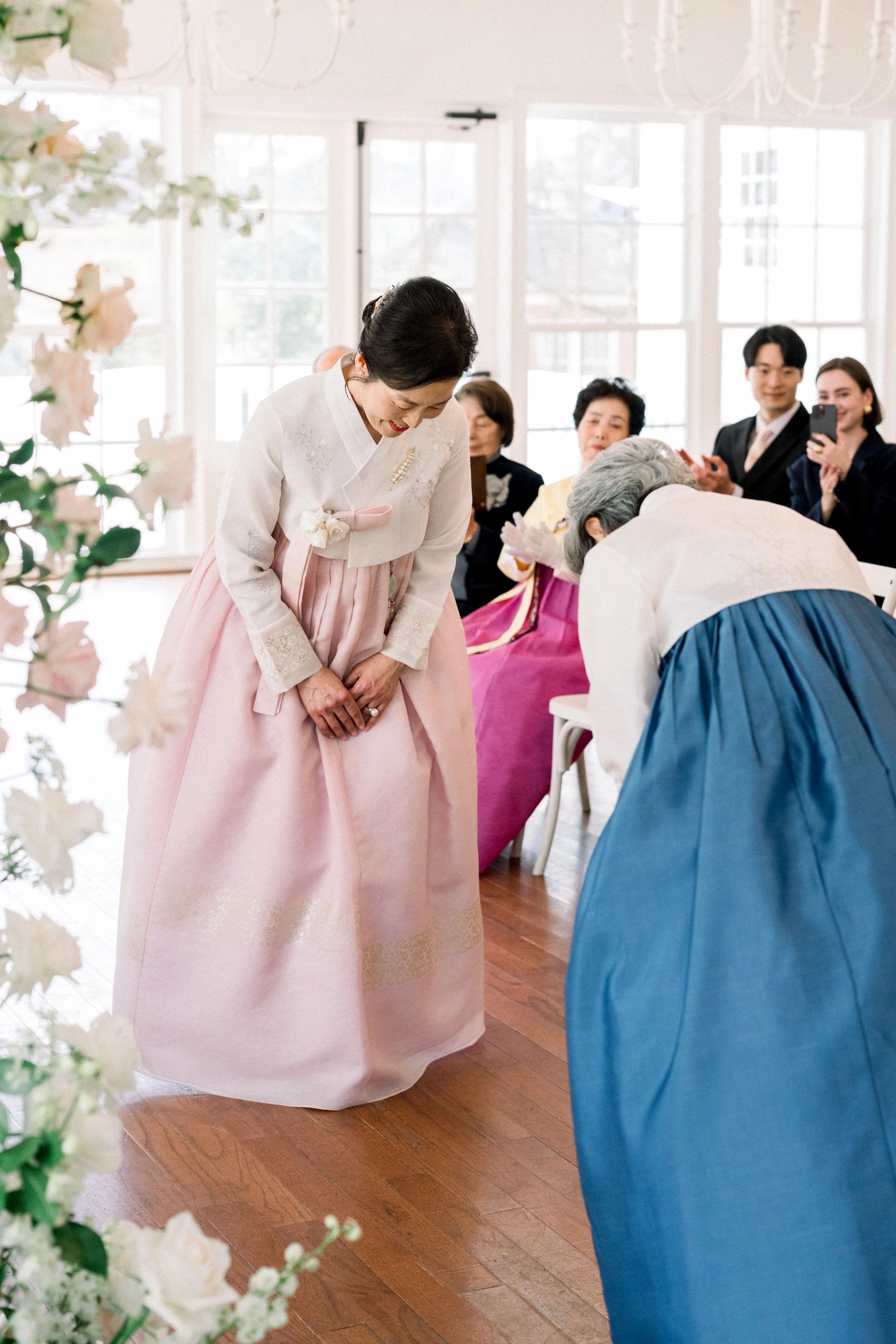 A woman in a pink traditional Korean hanbok bowing in front of an elderly woman at a gathering, with seated people watching and a smiling couple taking photos in the background.