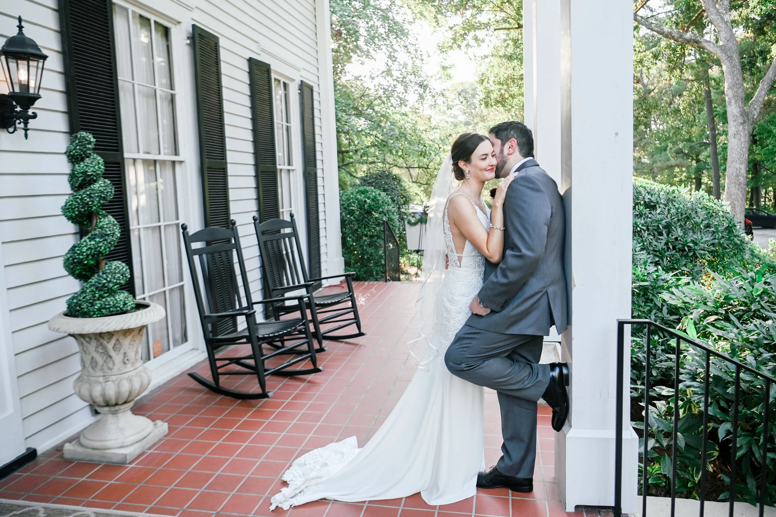A bride and groom share a tender moment on a porch, with the groom kneeling and kissing the bride on the cheek, while she smiles.