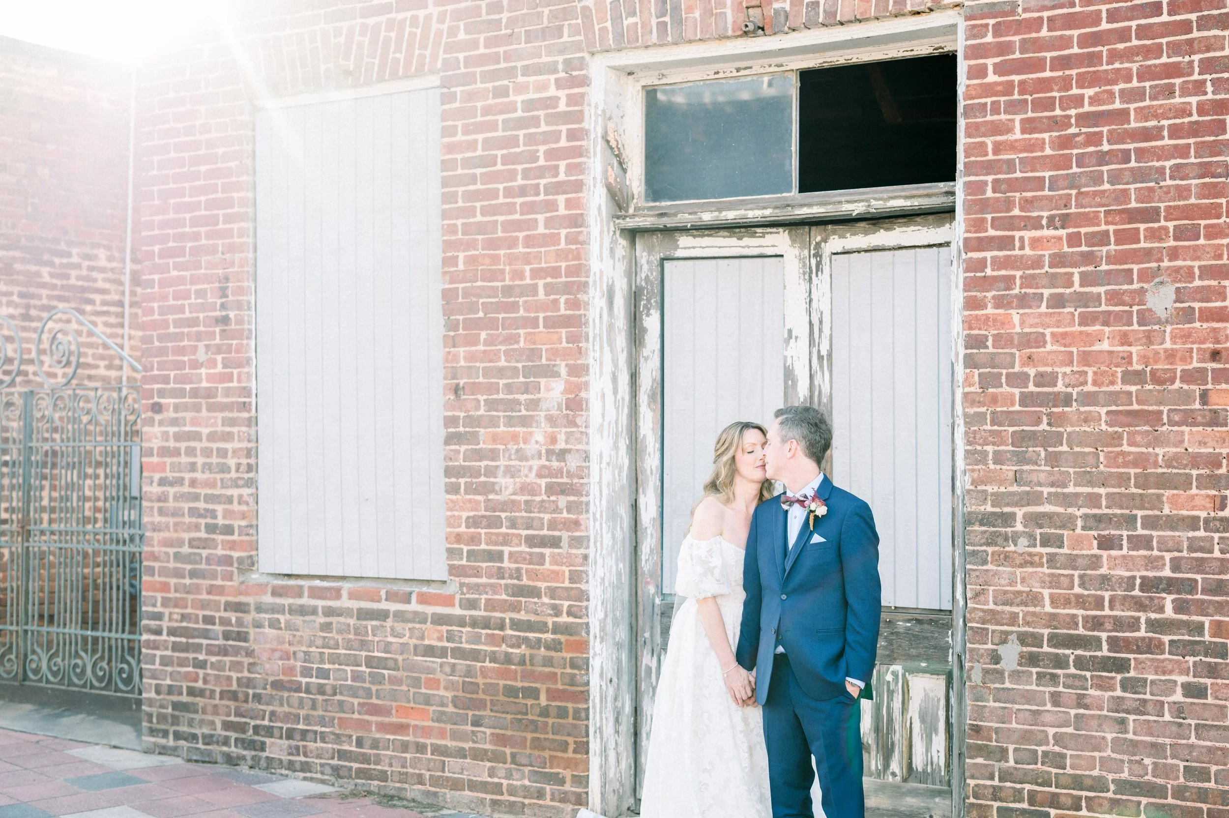 A bride and groom sharing a kiss, standing closely in front of a rustic brick building with a large window and weathered wooden door, during daytime.
