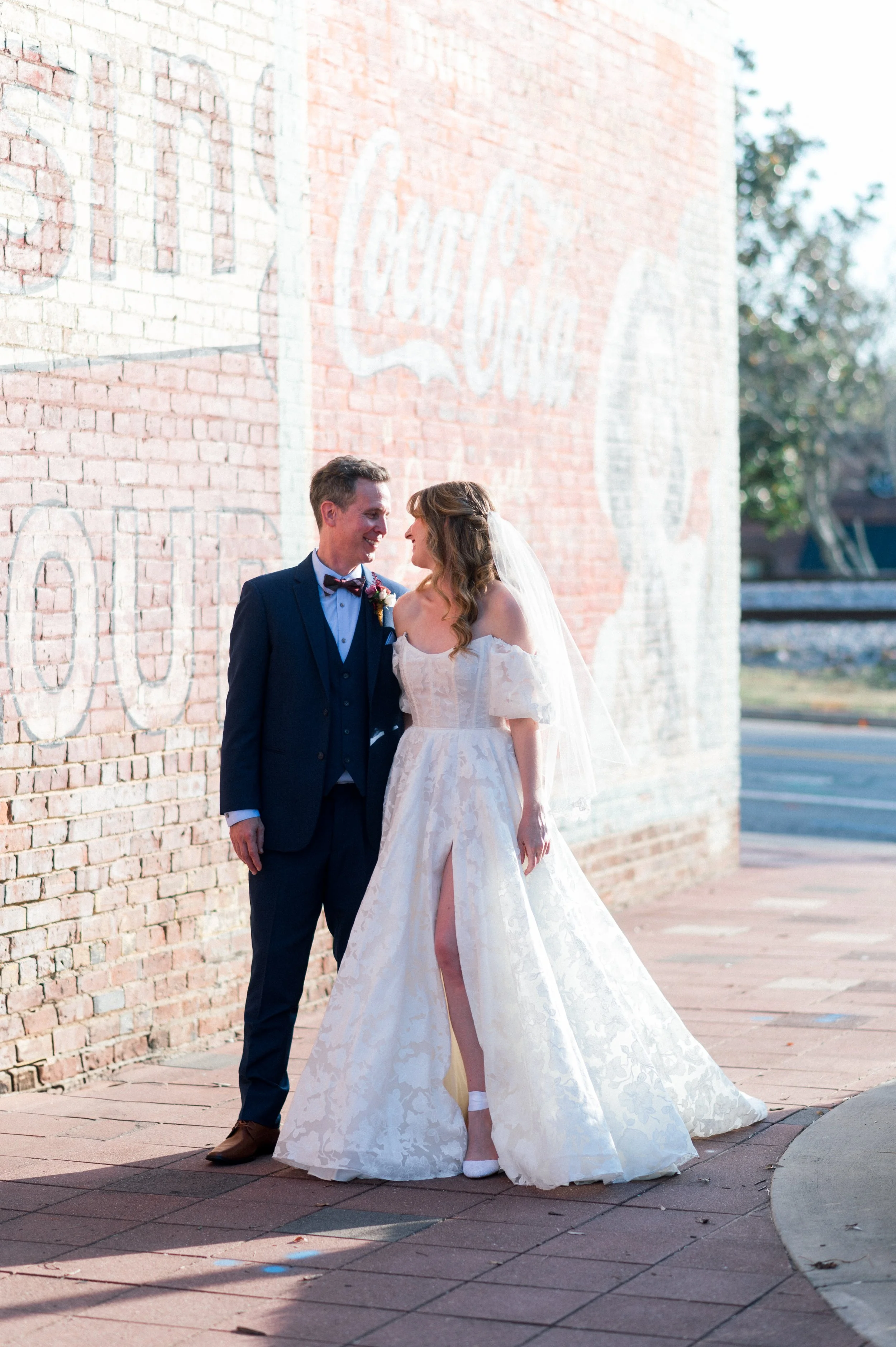 A newlywed couple standing close to each other outside near a brick wall with a faded Coca-Cola advertisement painted on it. The groom is wearing a dark suit with a bowtie and boutonniere. The bride is in a white wedding dress with a slit, wearing wh