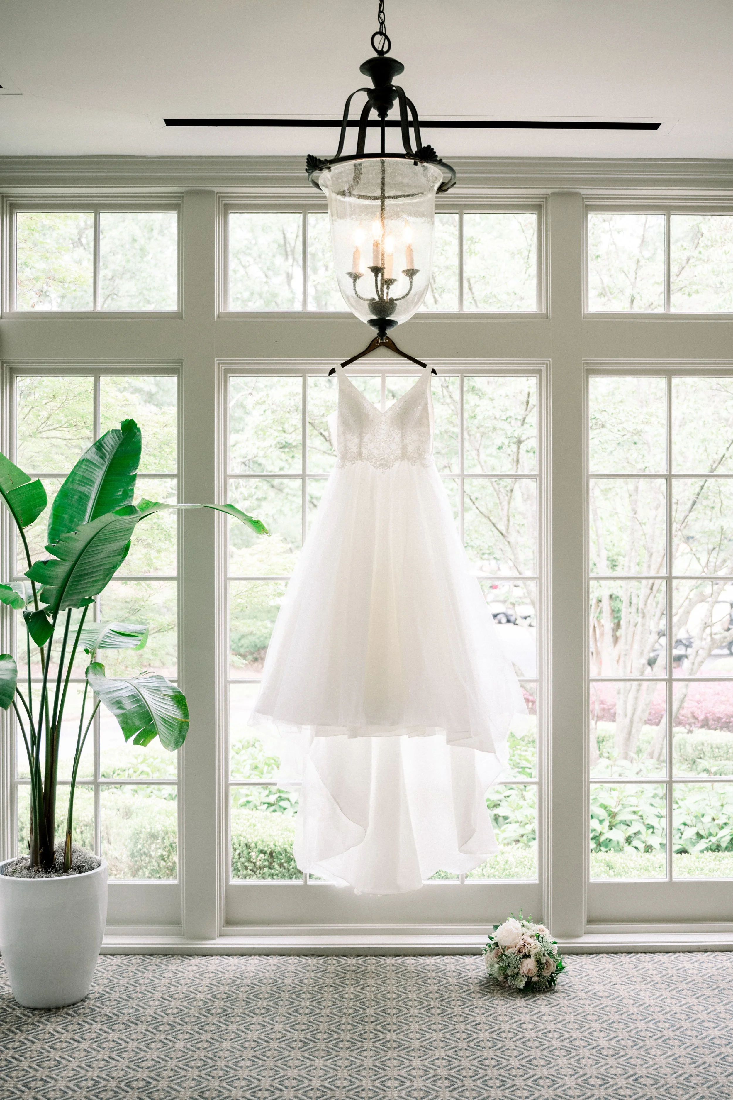 A wedding dress hanging from a hanger in front of large windows with trees outside. A potted plant is on the left, and a bouquet of flowers is on the floor in front of the window.