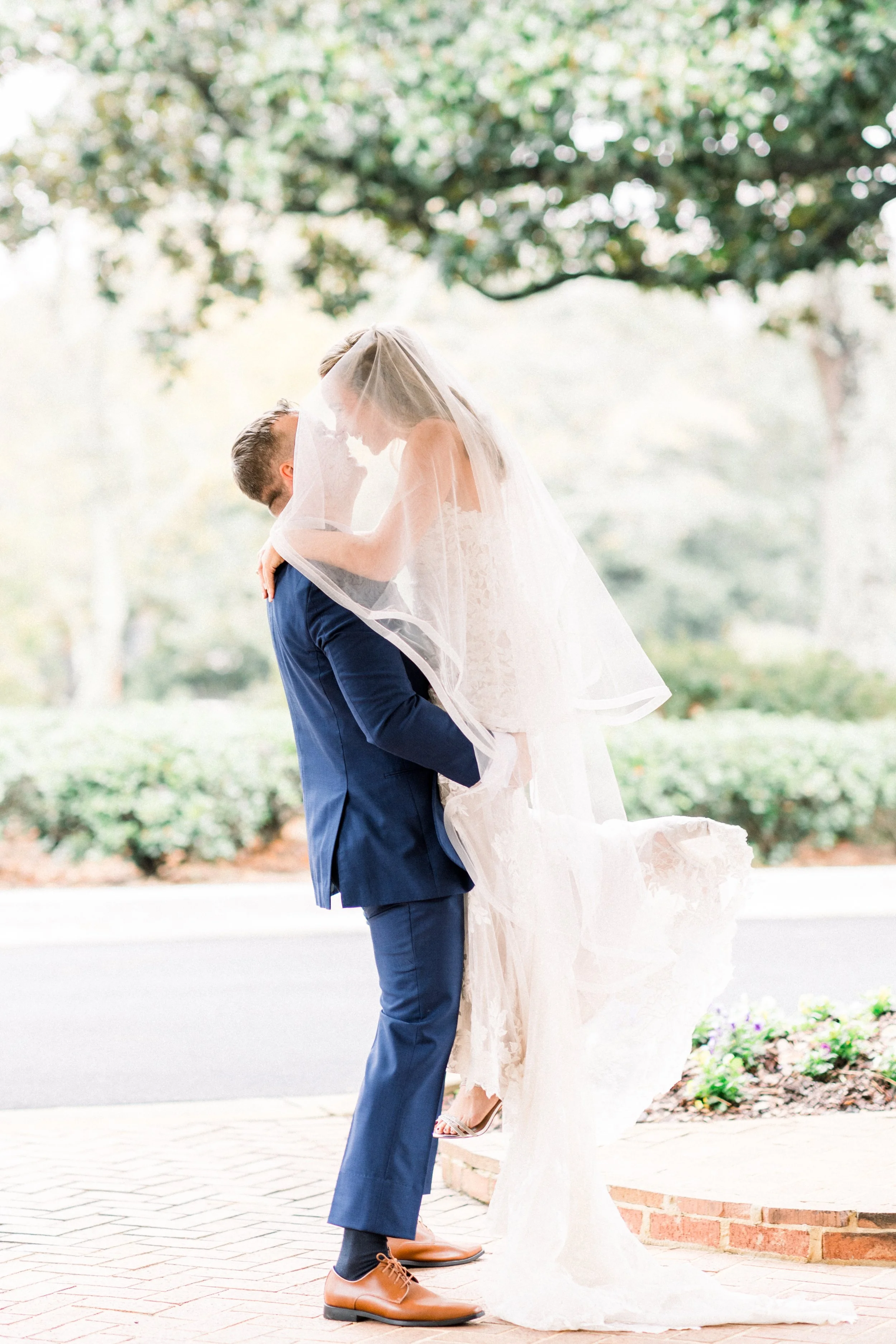 A couple dressed in wedding attire standing outdoors under a large tree, with the woman on the man's shoulders, kissing.