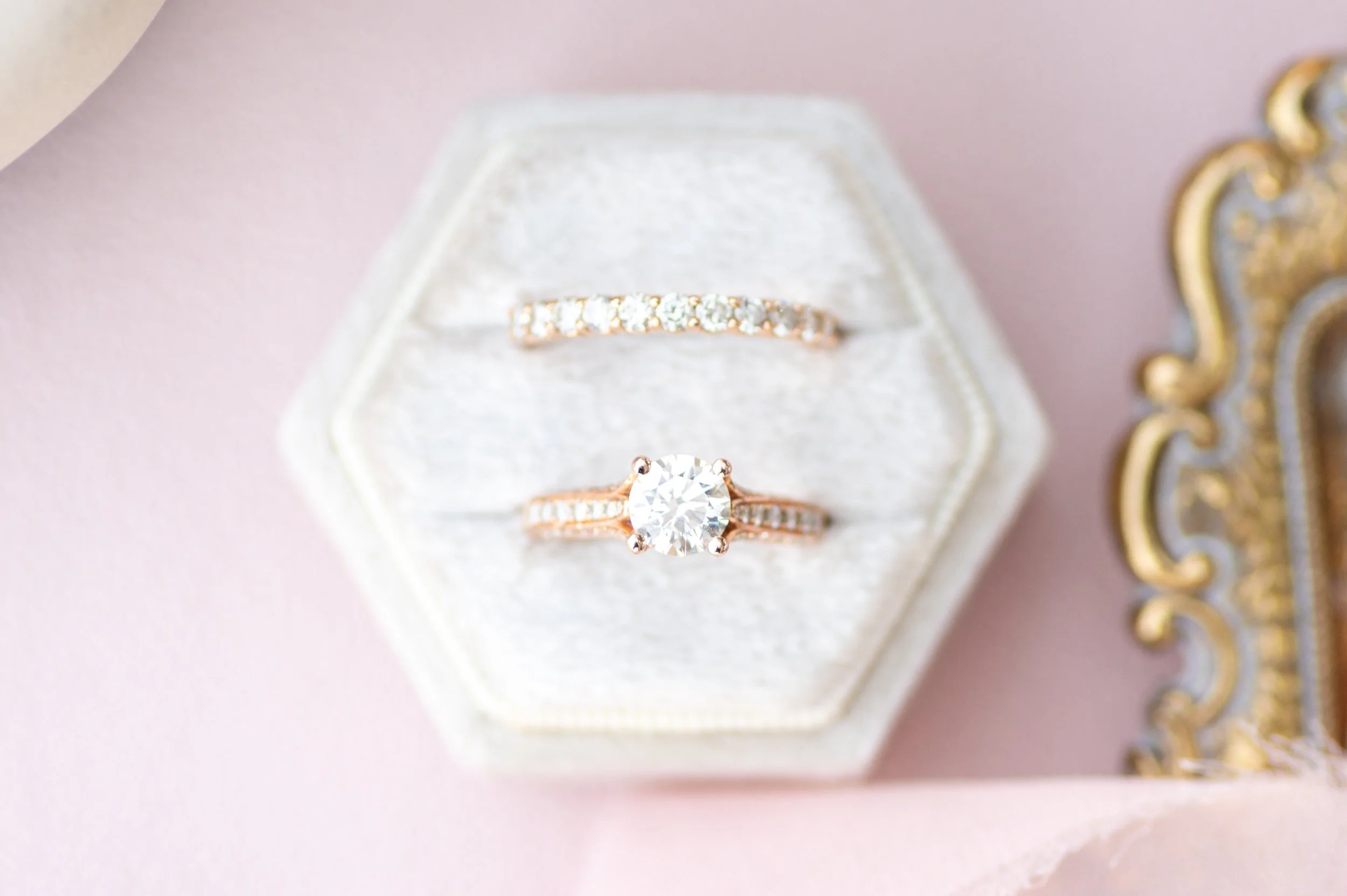 Close-up of a diamond engagement ring with a large round center stone, set in a rose gold band with small accent diamonds, placed inside a white jewelry box with a textured surface, on a pink background.