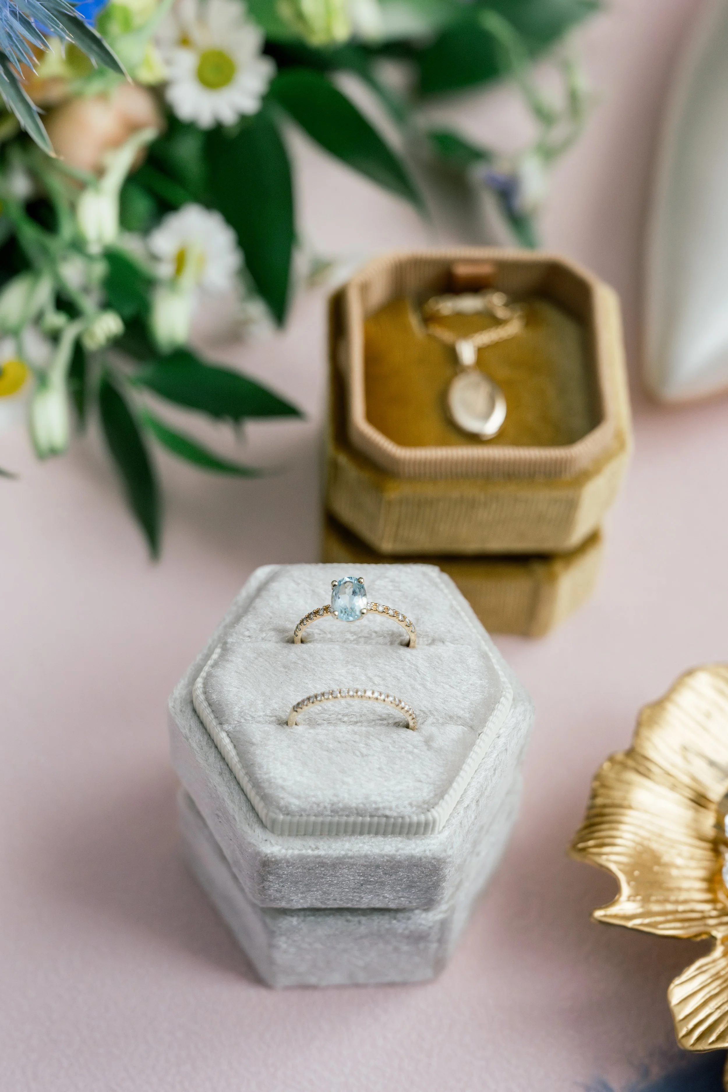 A close-up of a jewelry display featuring a silver ring with a large oval-shaped blue gemstone and small surrounding diamonds, placed on a white plush jewelry box. In the background, a small hexagonal jewelry box with a gold chain necklace containing