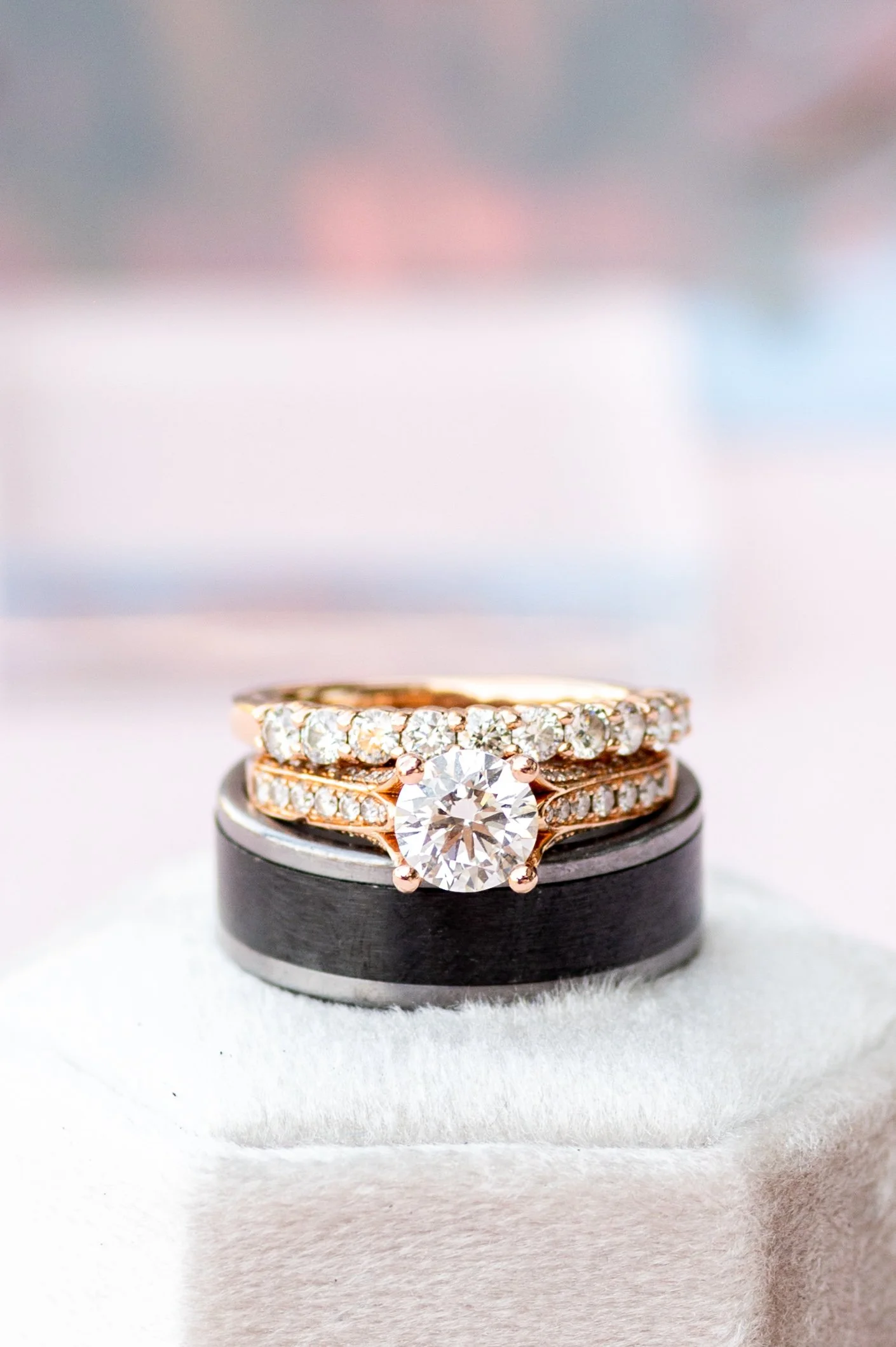 Close-up of a diamond engagement ring with a large round center stone and diamond-studded band, displayed on a black ring holder with a blurred pastel background.