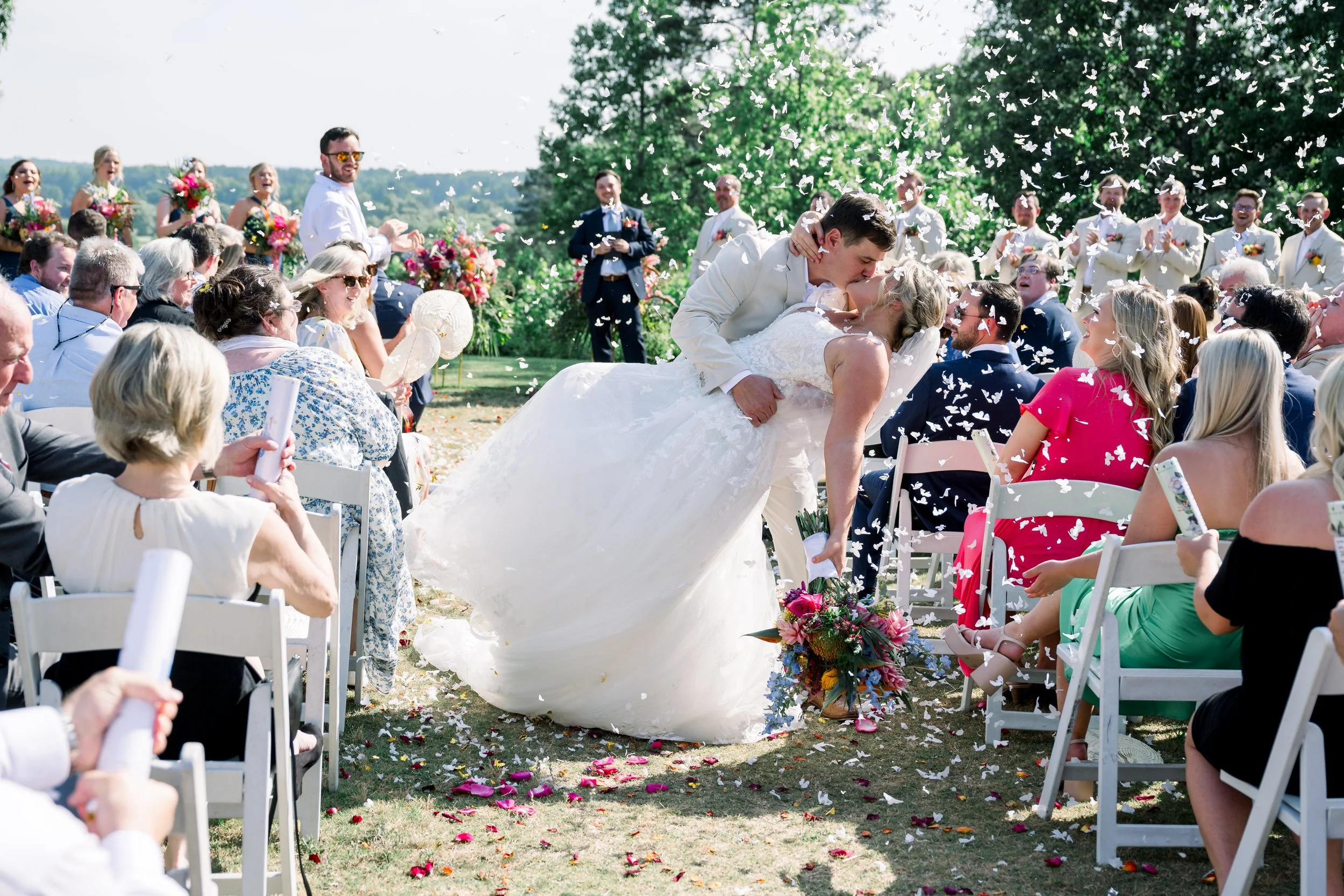 A newlywed couple sharing a kiss during an outdoor wedding ceremony, surrounded by seated guests, with flower petals falling from above and lush green trees in the background.