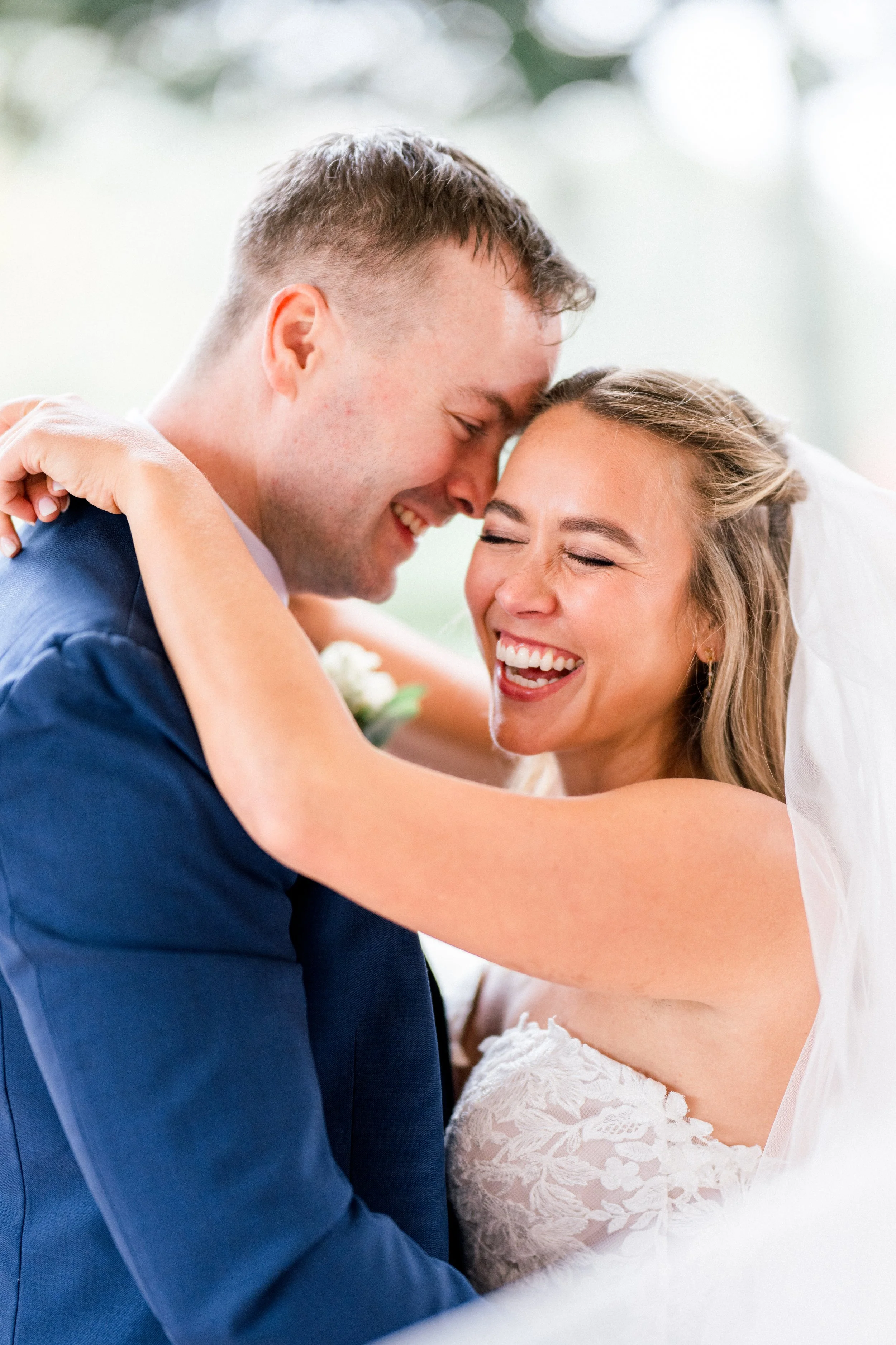 A happy bride and groom are embracing each other, with their foreheads touching and smiling with their eyes closed during their wedding celebration.