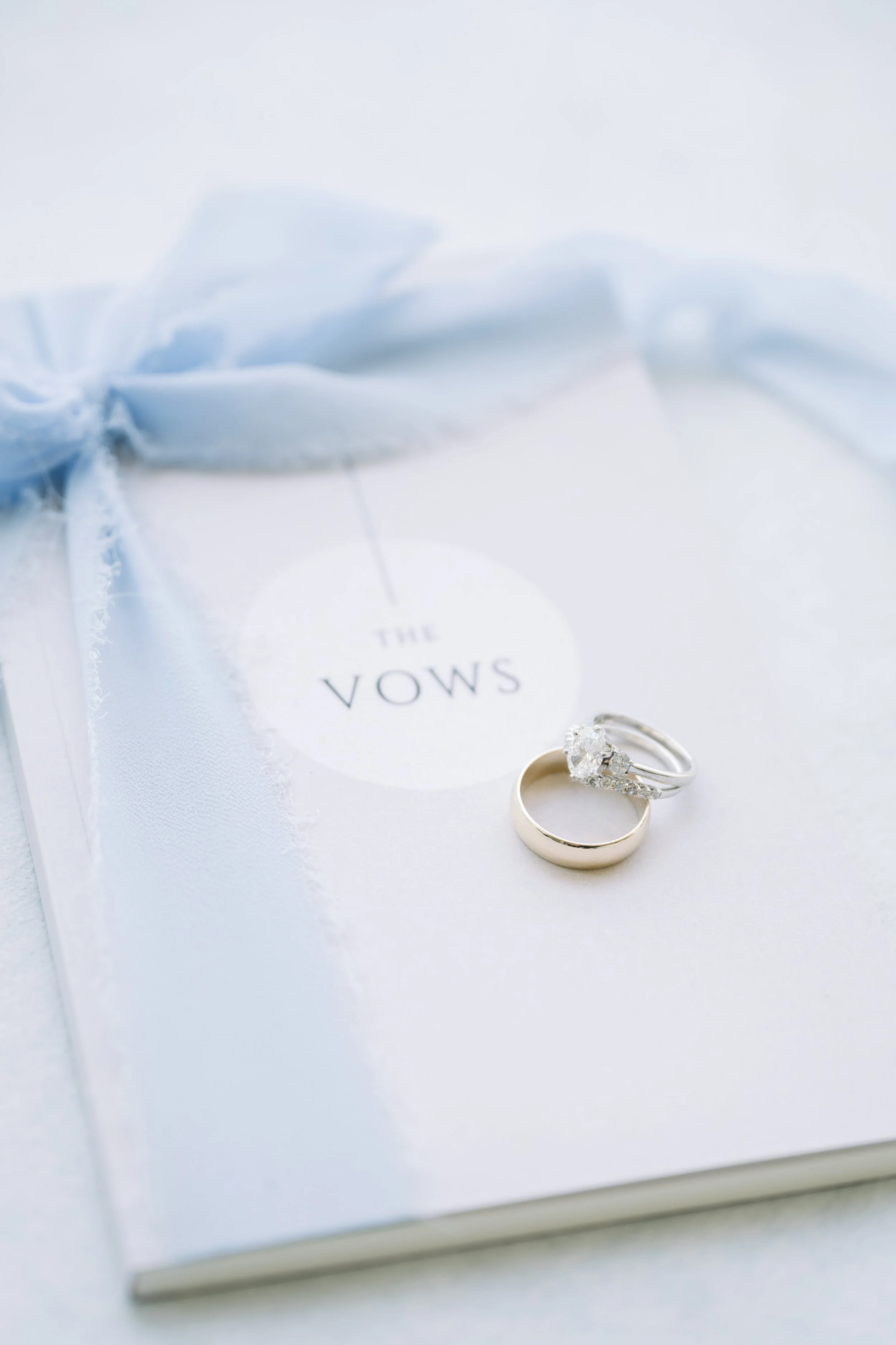 Close-up of wedding rings on a white surface with a blurred wedding vow booklet in the background.