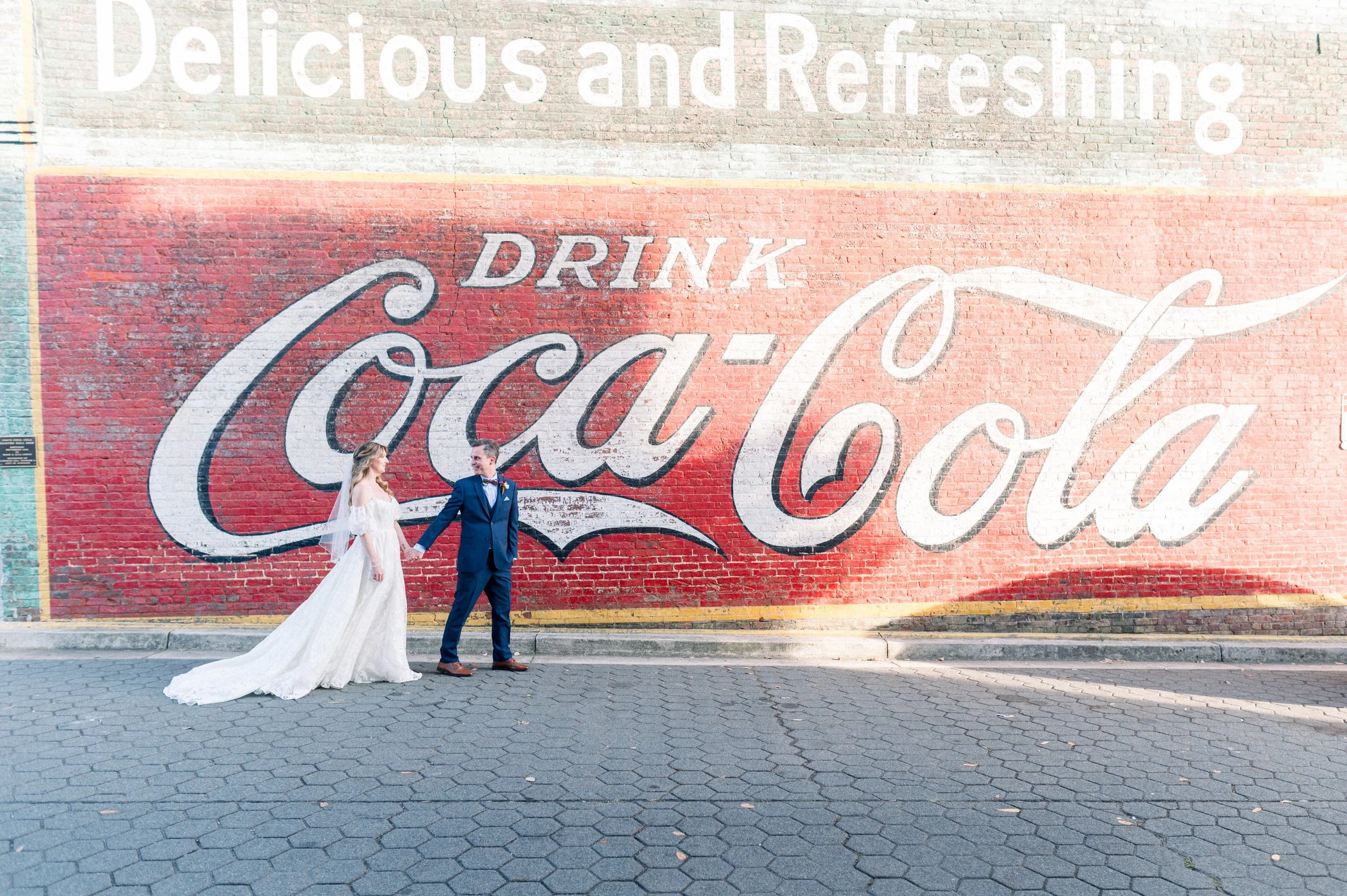 A bride and groom holding hands and smiling while standing in front of a large Coca-Cola advertisement painted on a brick wall, on a city sidewalk.