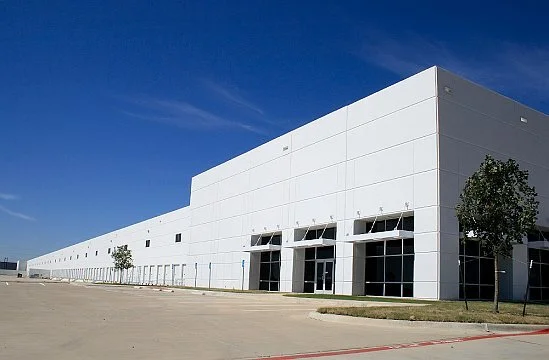 Large white warehouse with multiple loading bays and trees in front, under a clear blue sky.