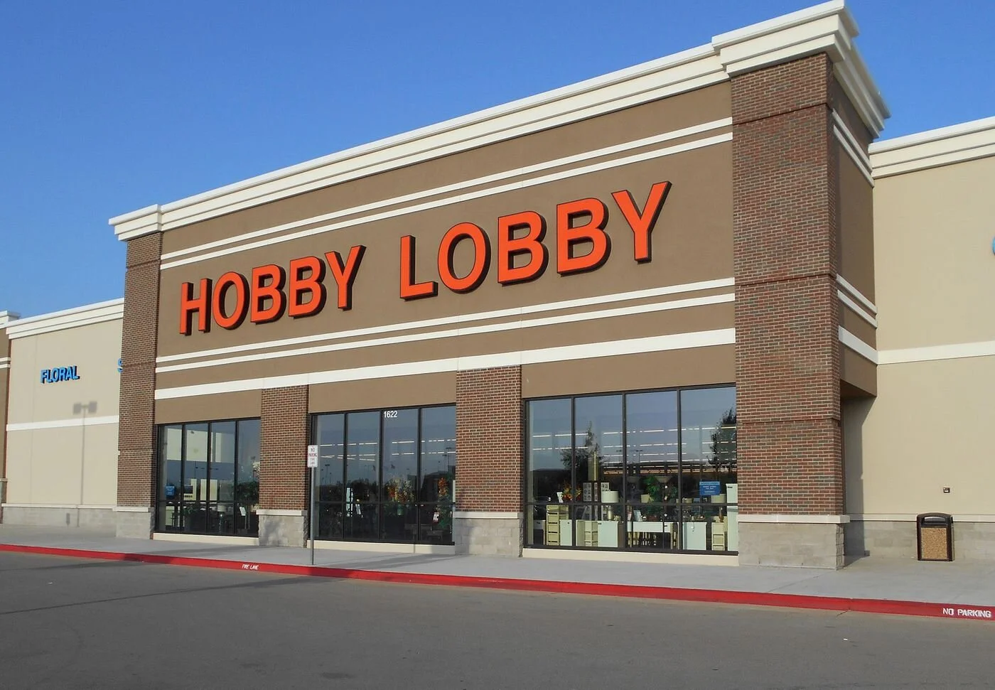 Exterior of a retail store with a large sign reading 'HOBBY LOBBY' in orange letters, featuring large glass windows and brick accents at the entrance.