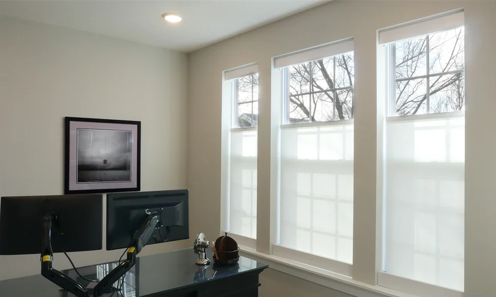 Modern office with two computer monitors on a desk, framed black and white artwork on the wall, and three large windows letting in natural light.
