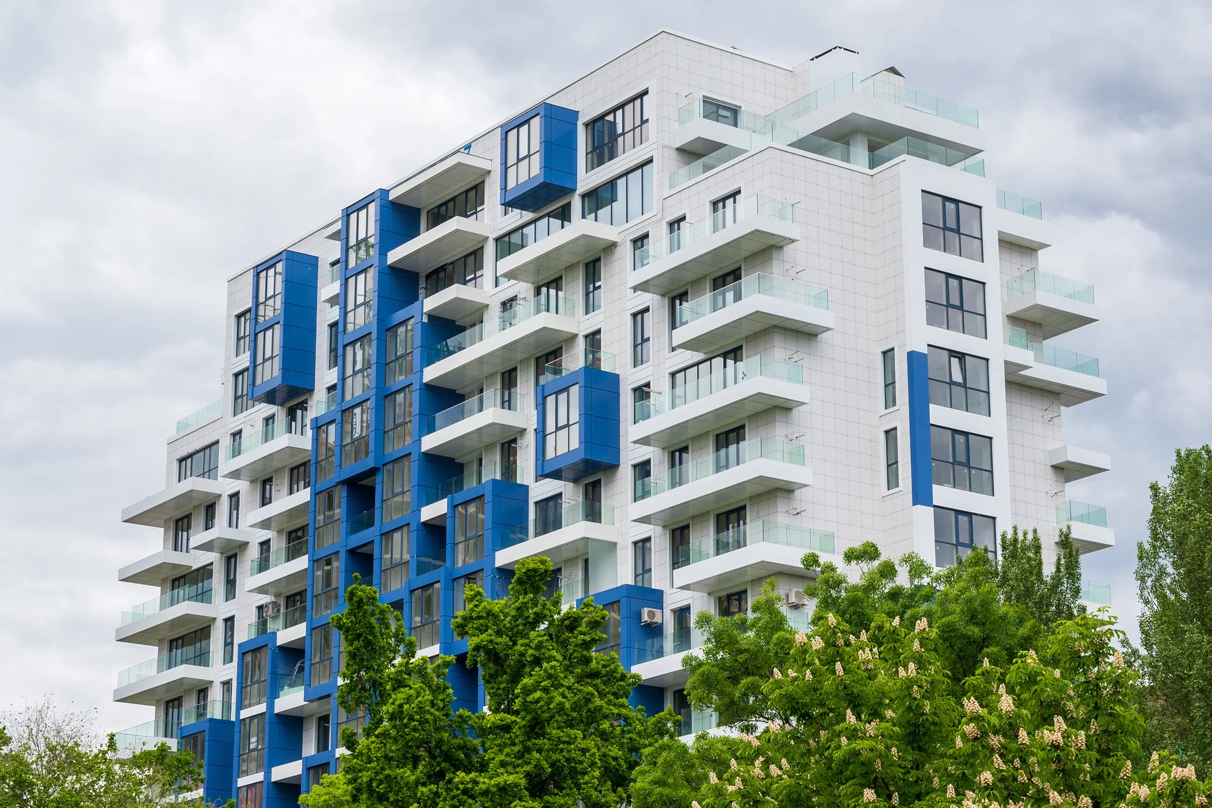 A modern multi-story apartment building with a white and blue facade, balconies, and large windows, surrounded by trees under a cloudy sky.