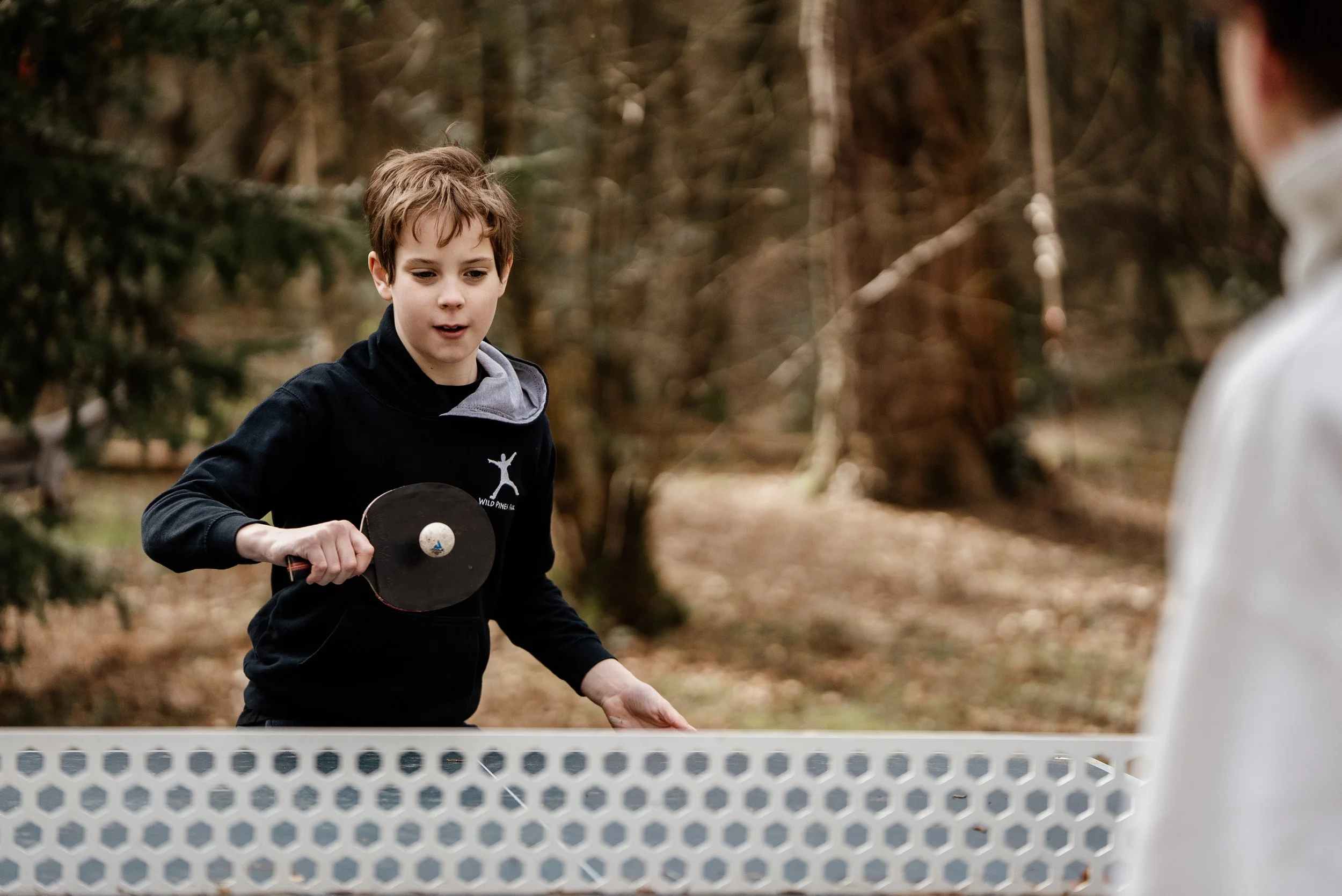 Young boy playing pickleball outdoors in a wooded area, holding a paddle with a ball on it, wearing a black hoodie, with a woman partially visible in the background.