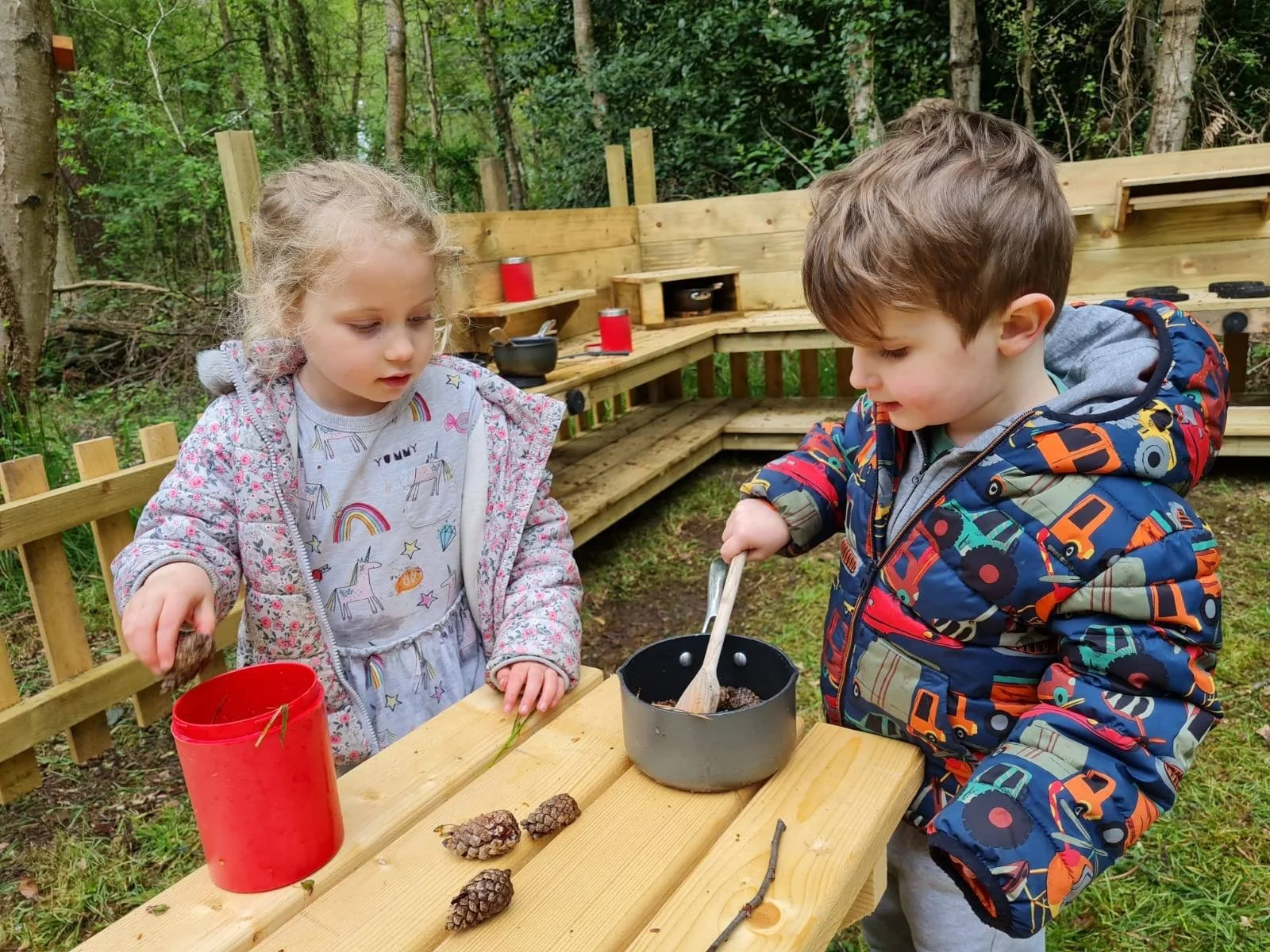 Two young children, a girl and a boy, engaged in a craft activity outdoors at a wooden table in a forested area. The girl is holding a pine cone, and the boy is stirring a mixture in a small pot. There are pine cones and a red container on the table.