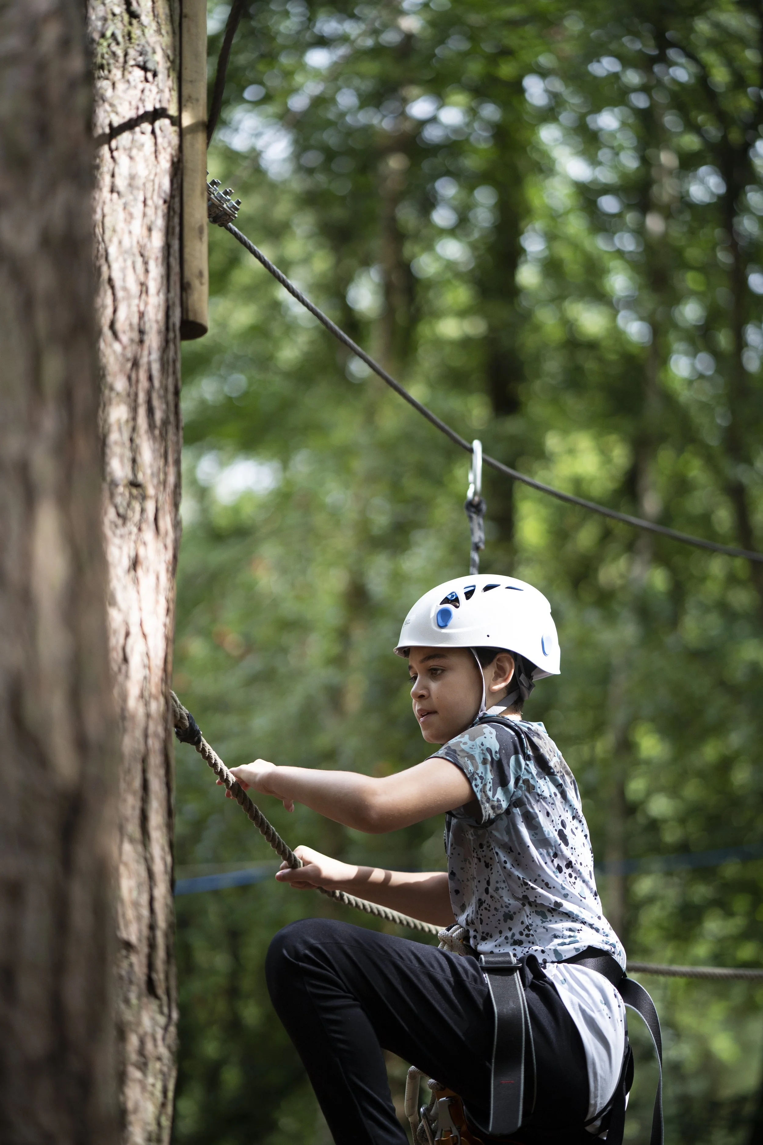 A young boy wearing a helmet and camouflage shirt climbing a rope on a tree in a forest setting, with a focus on the boy and the tree trunk.