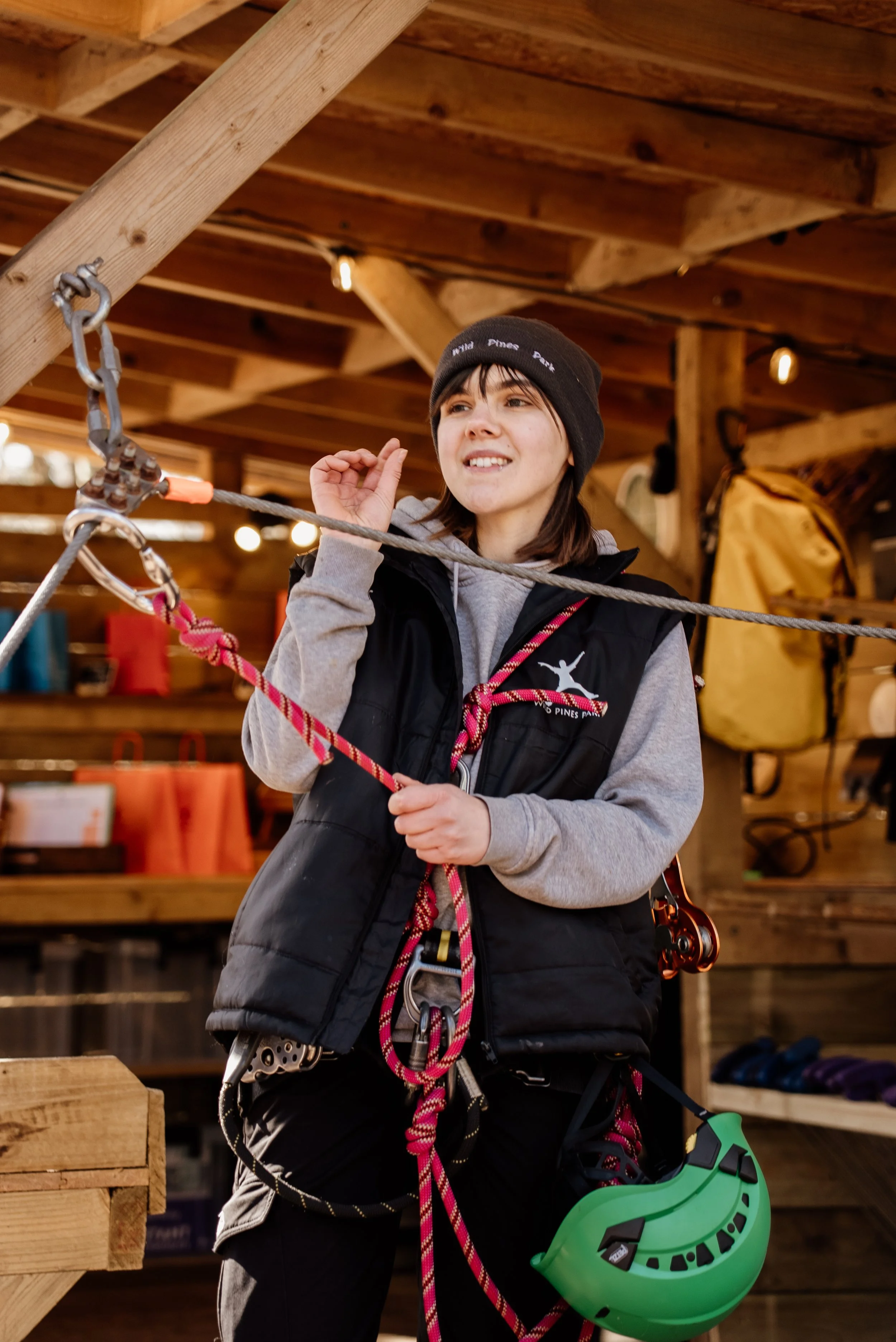 A young woman wearing a black beanie, gray hoodie, and black vest, with climbing gear, stands inside a wooden cabin or climbing gym, smiling and adjusting her hair.