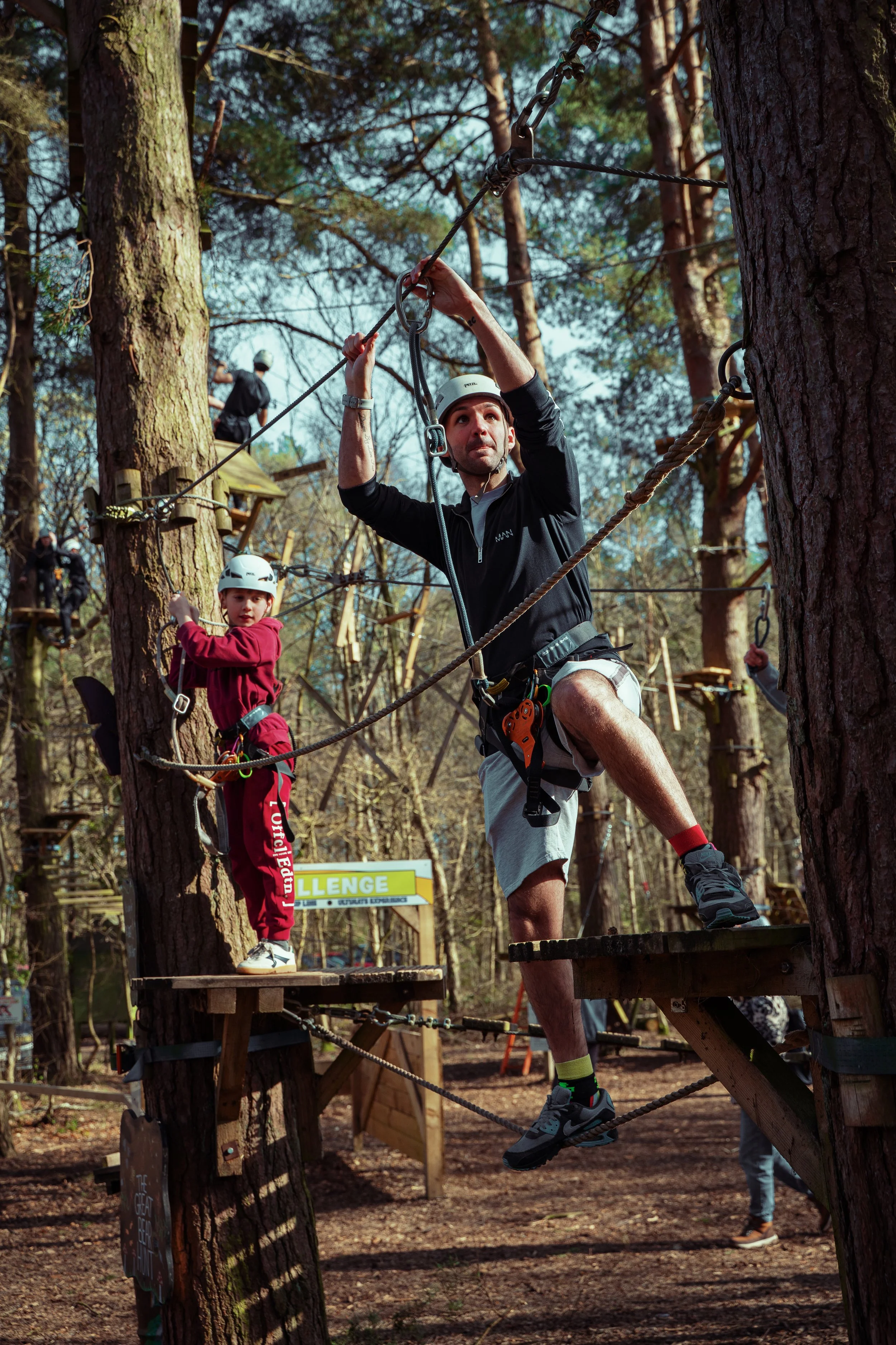 Man and children on a ropes course in a forest, wearing helmets and harnesses, climbing platforms connected by ropes.