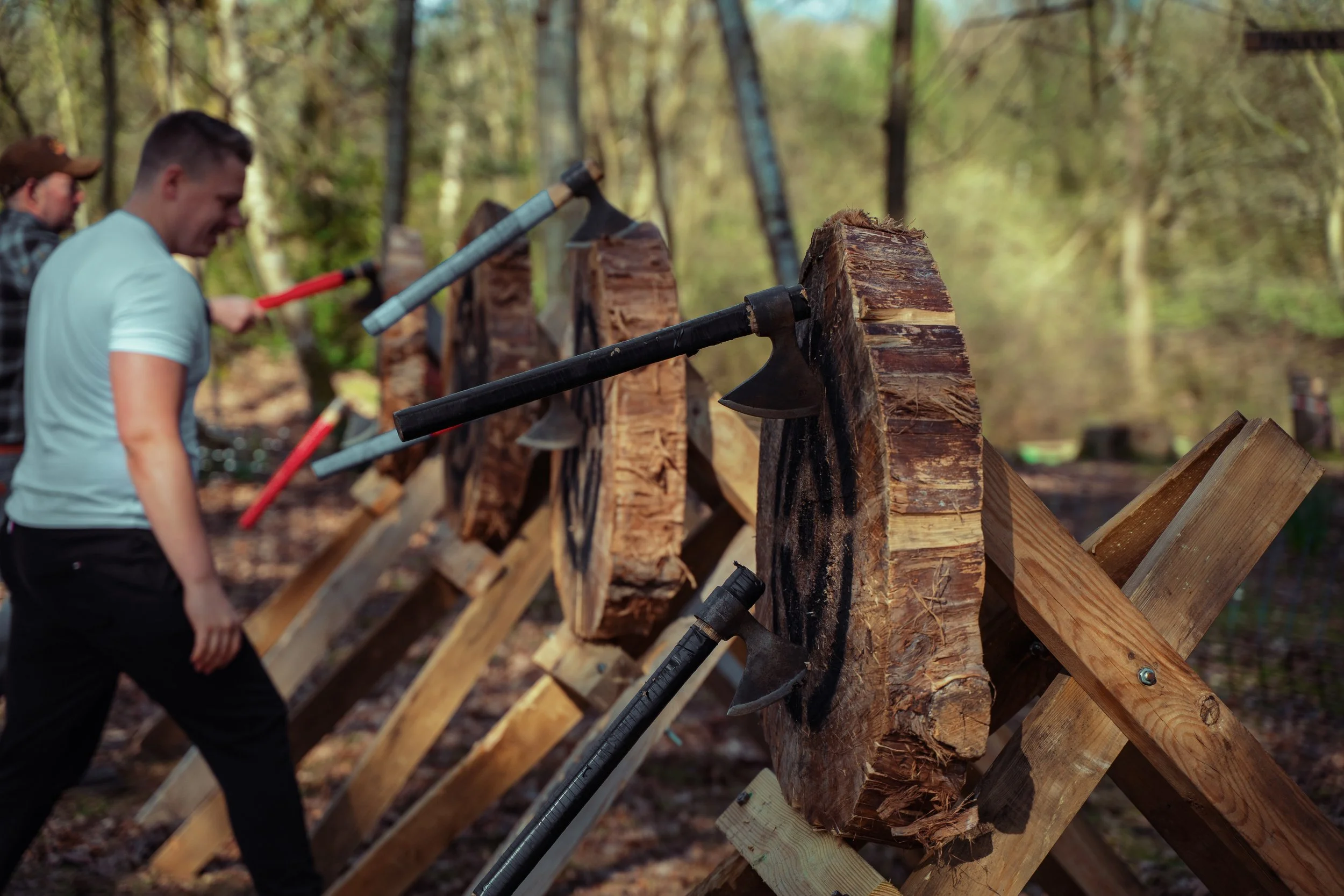 People chopping logs with axes in a wooded outdoor setting.