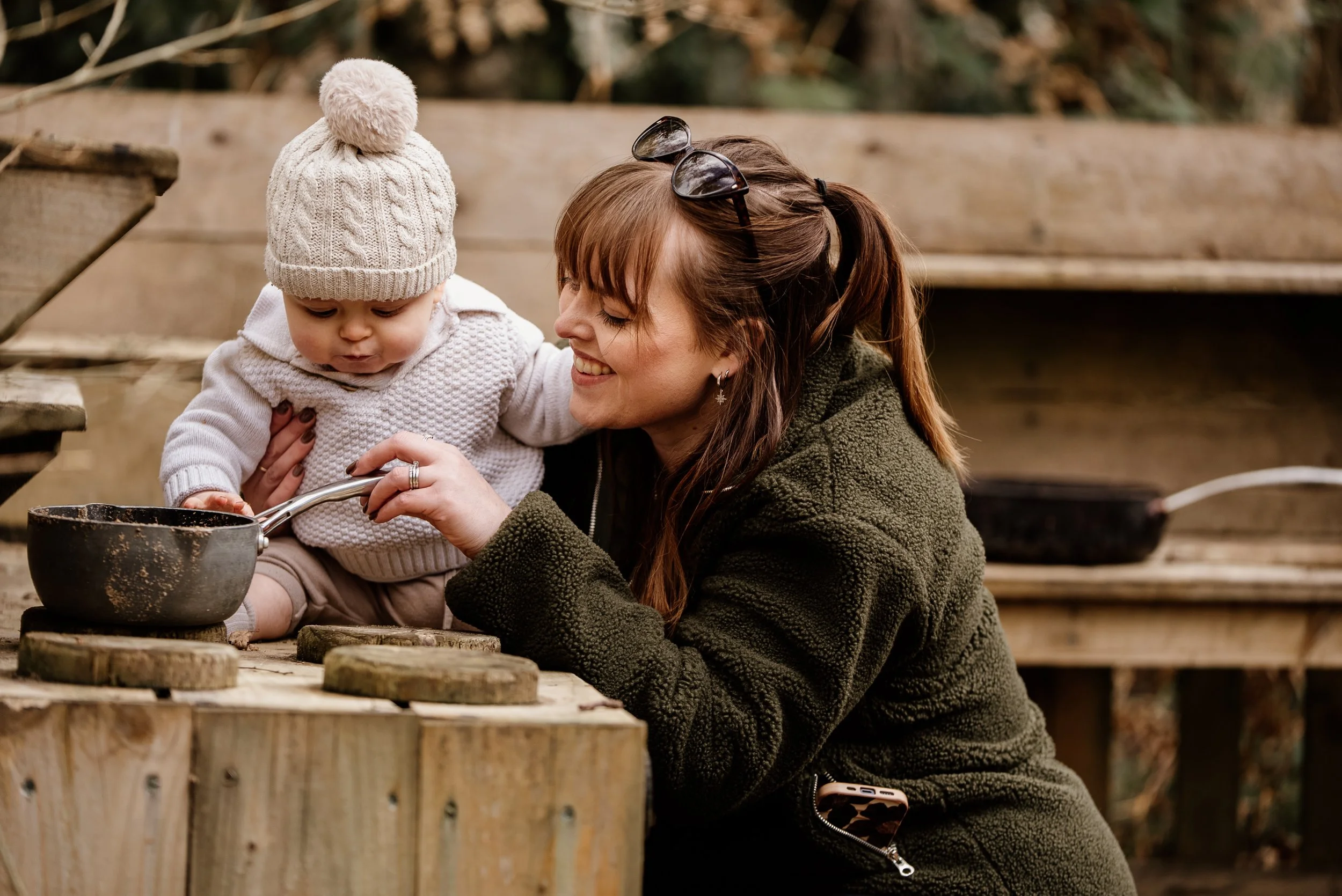 A woman and a young child outdoors on a wooden platform, with the woman smiling and the child looking down at a pot they are holding together.