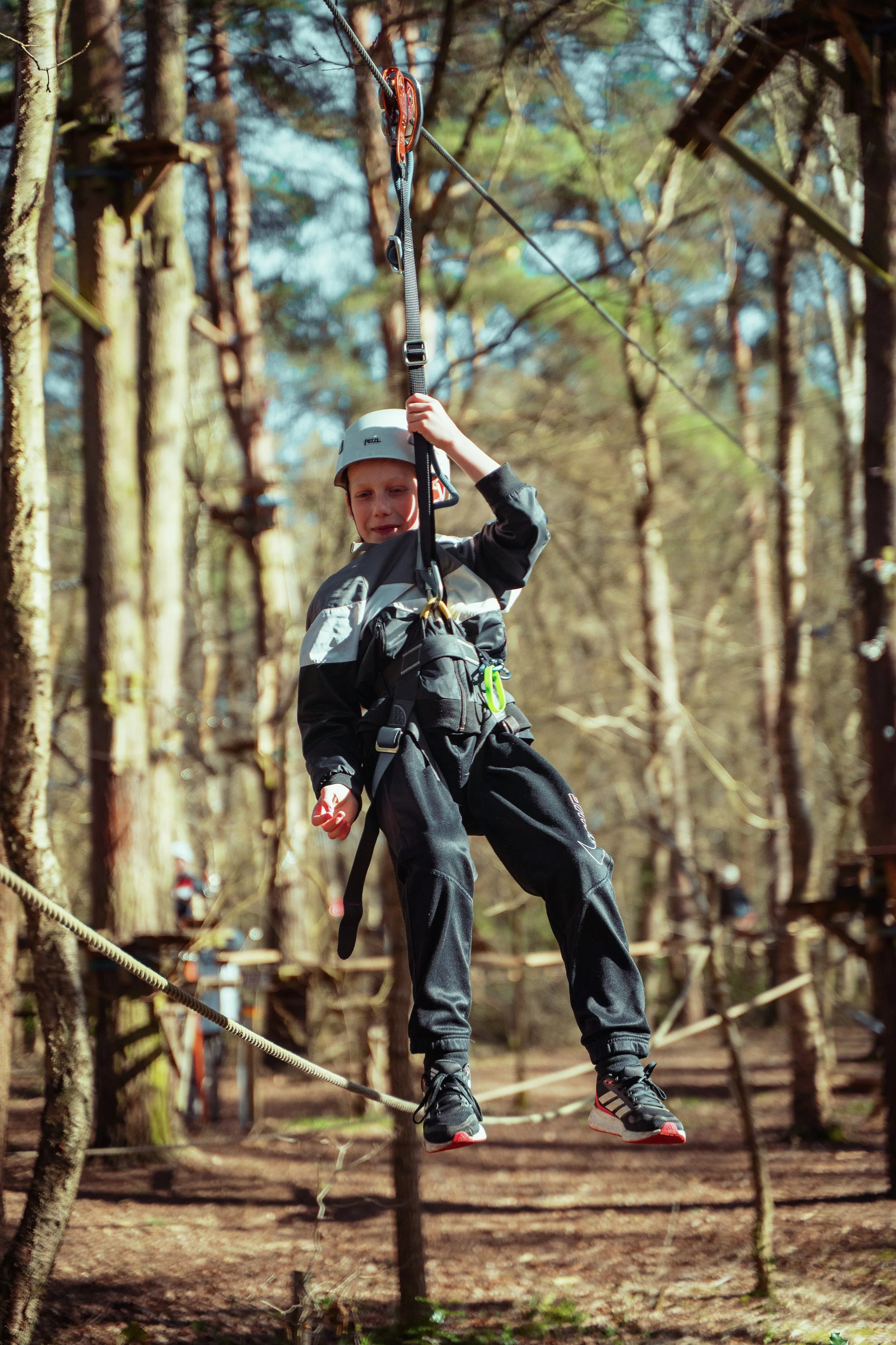 A young boy wearing a helmet and harness zip-lining through a forest with tall trees and a wooden platform in the background.