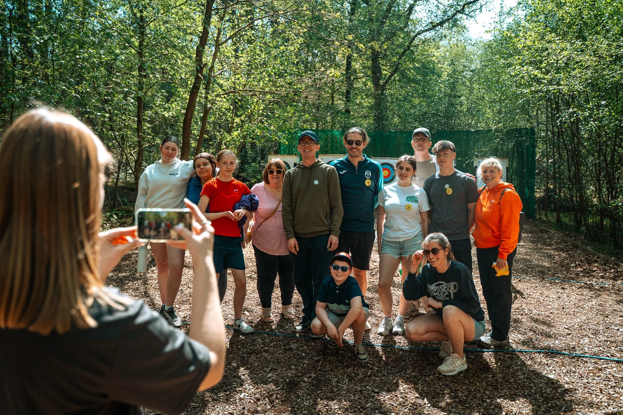 Group of people, including children and adults, posing for a photo in a wooded outdoor setting during daytime.