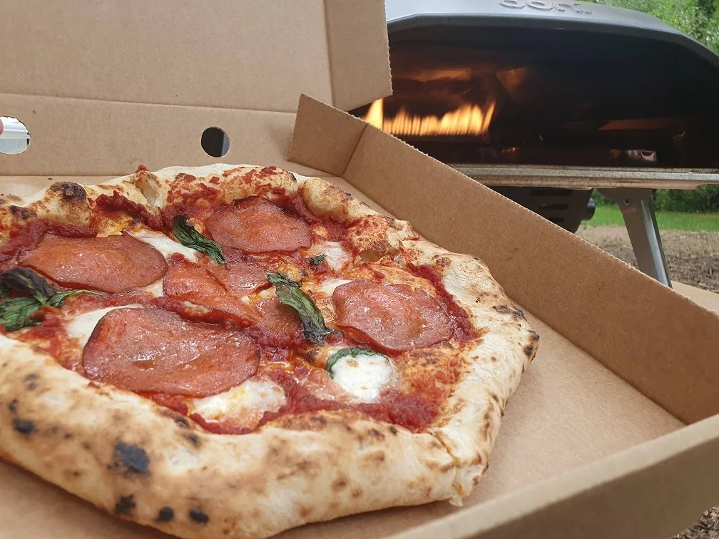 A pepperoni pizza with basil leaves in an open pizza box, with a pizza oven in the background.