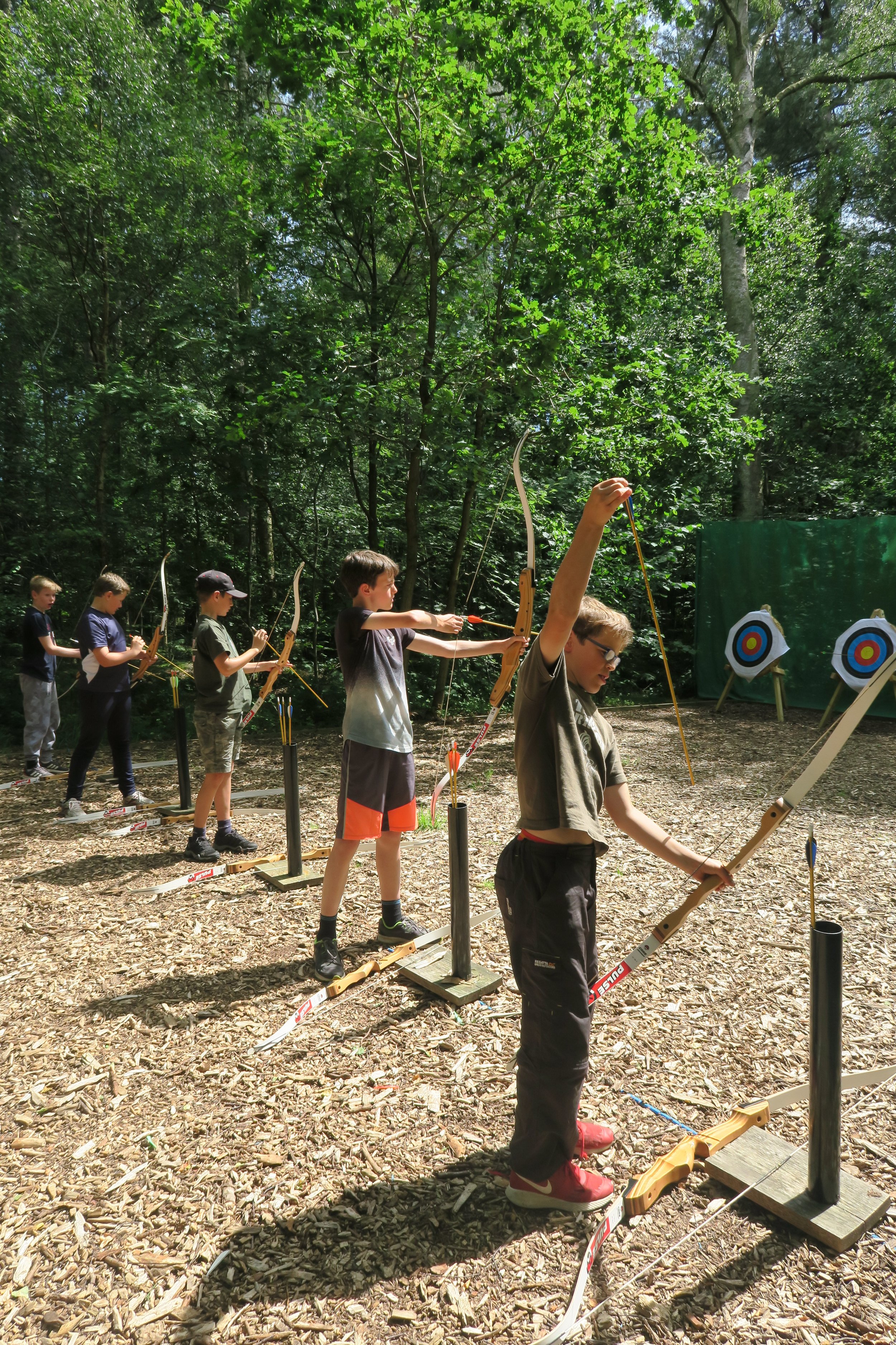 A group of children participating in archery in a wooded outdoor area, standing in a line and aiming bows at targets.