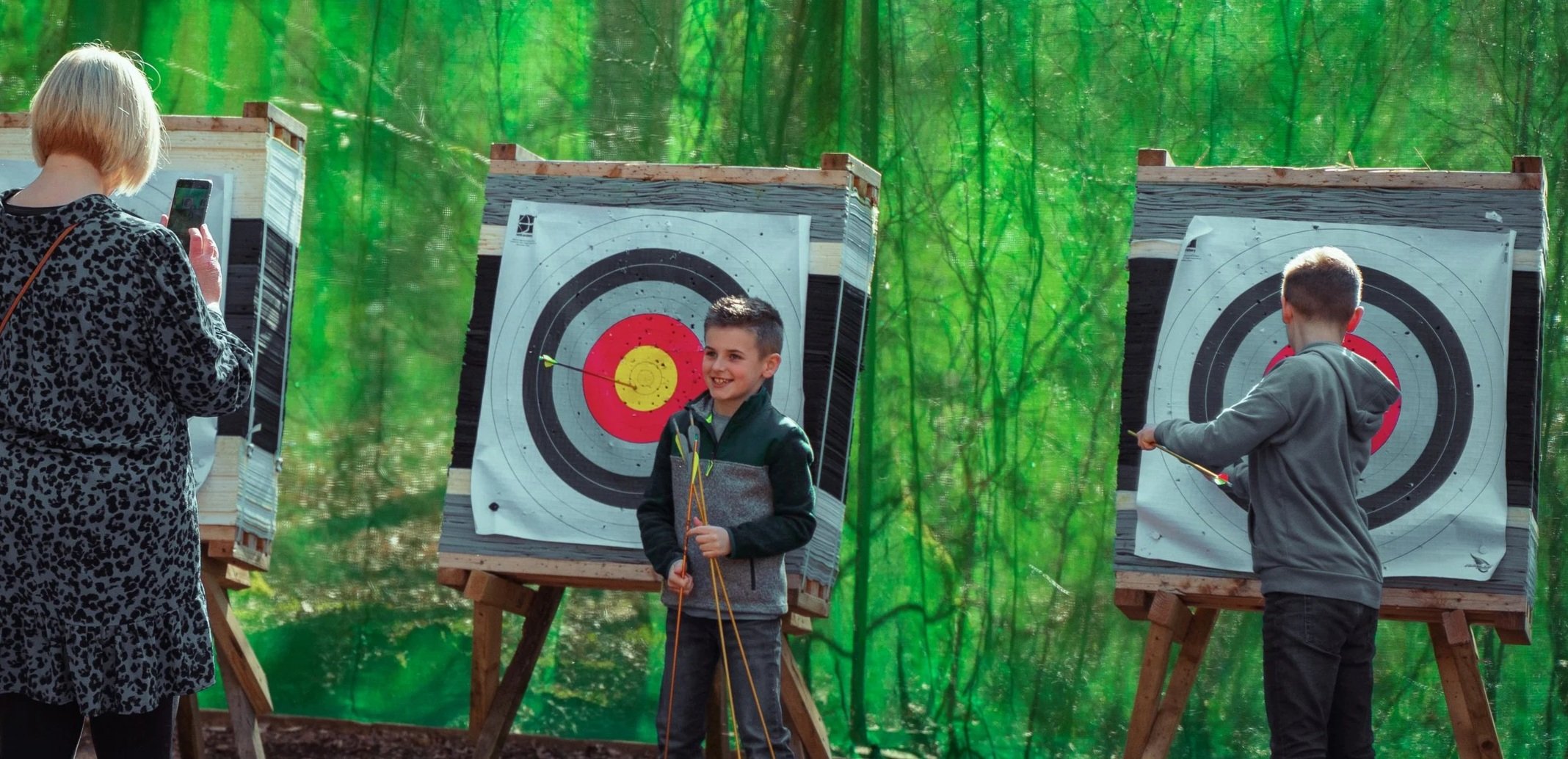 Two boys aiming bows and arrows at target boards in a wooded outdoor setting, with a woman taking a photo of them.
