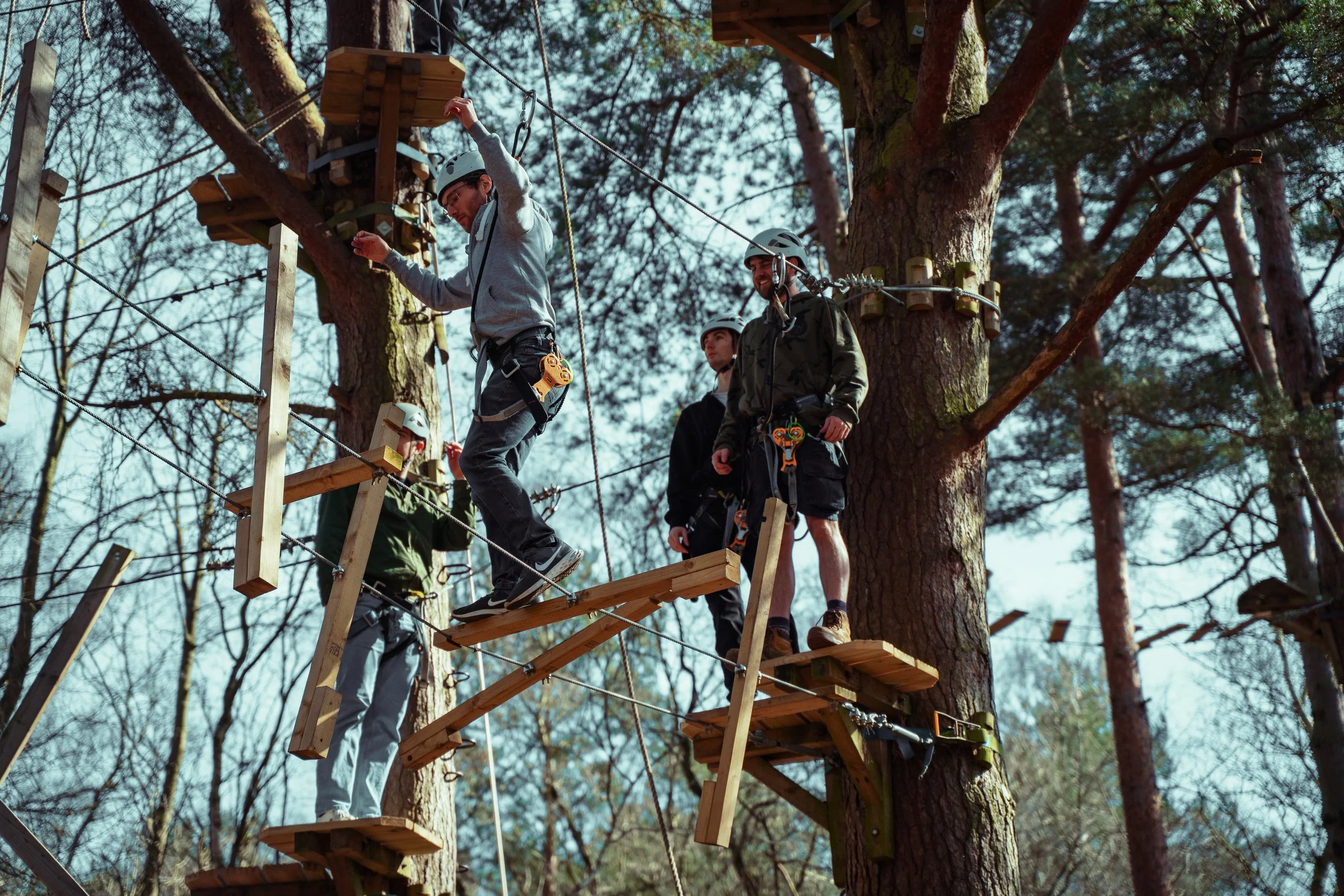 A group of people wearing helmets walking on a wooden obstacle course in a forest, with trees and sky in the background.