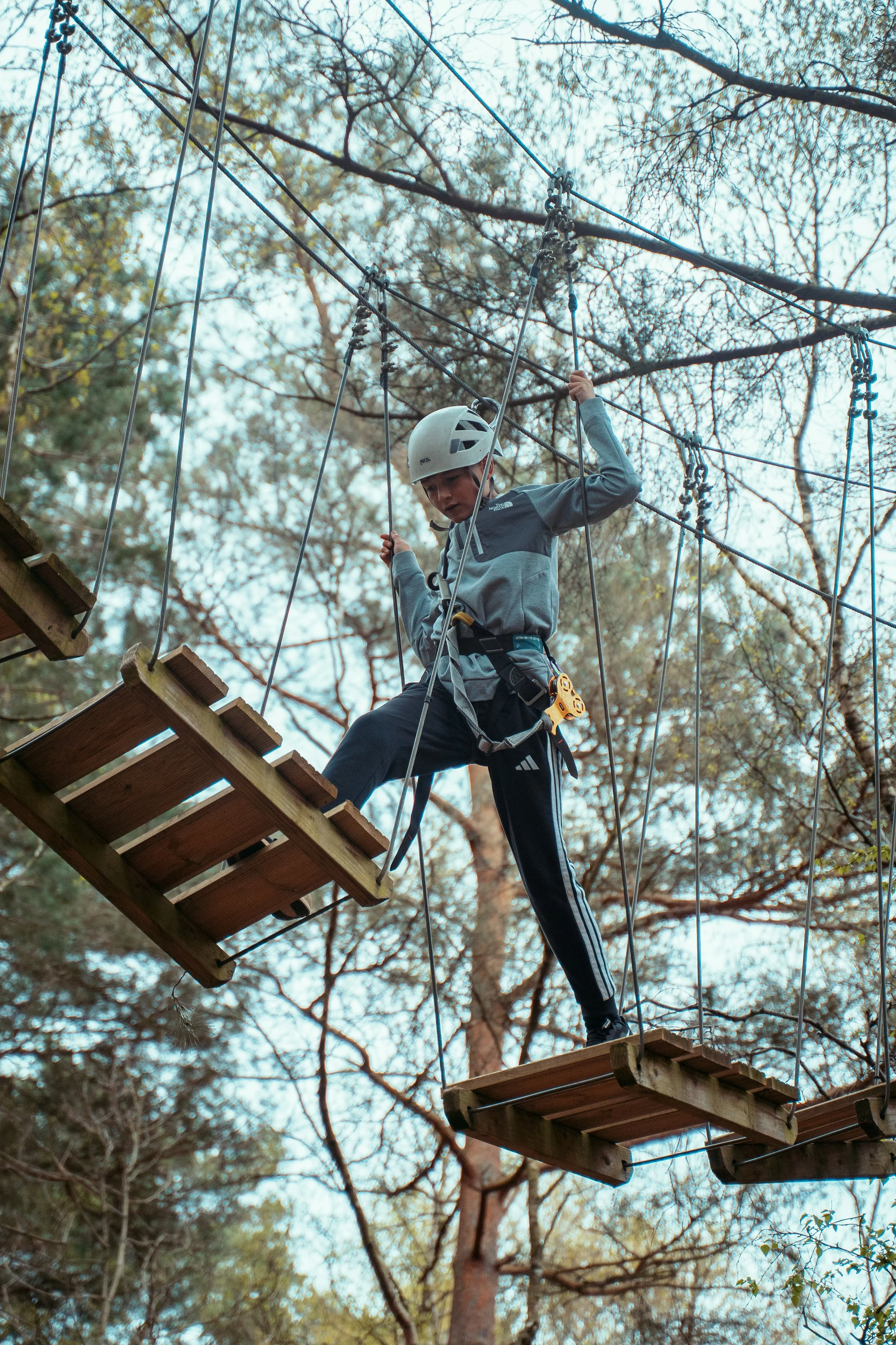 A boy in a helmet and harness crossing a wooden bridge in a treetop adventure course with trees and sky in the background.