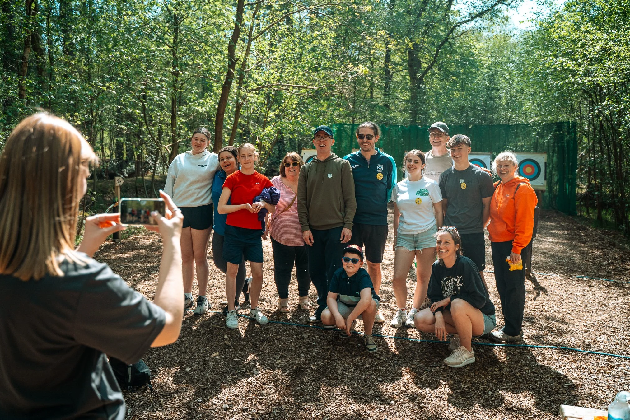 A group of people posing for a photo in a wooded outdoor area, some wearing sunglasses and casual clothing, while a woman in the foreground is taking the picture with her phone.
