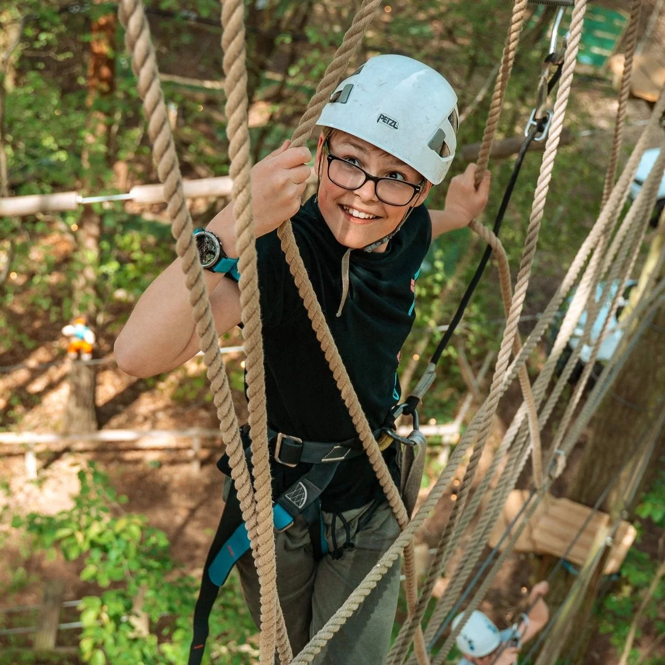 Young boy with glasses wearing a white helmet and harness, climbing on a rope bridge in a forest setting.