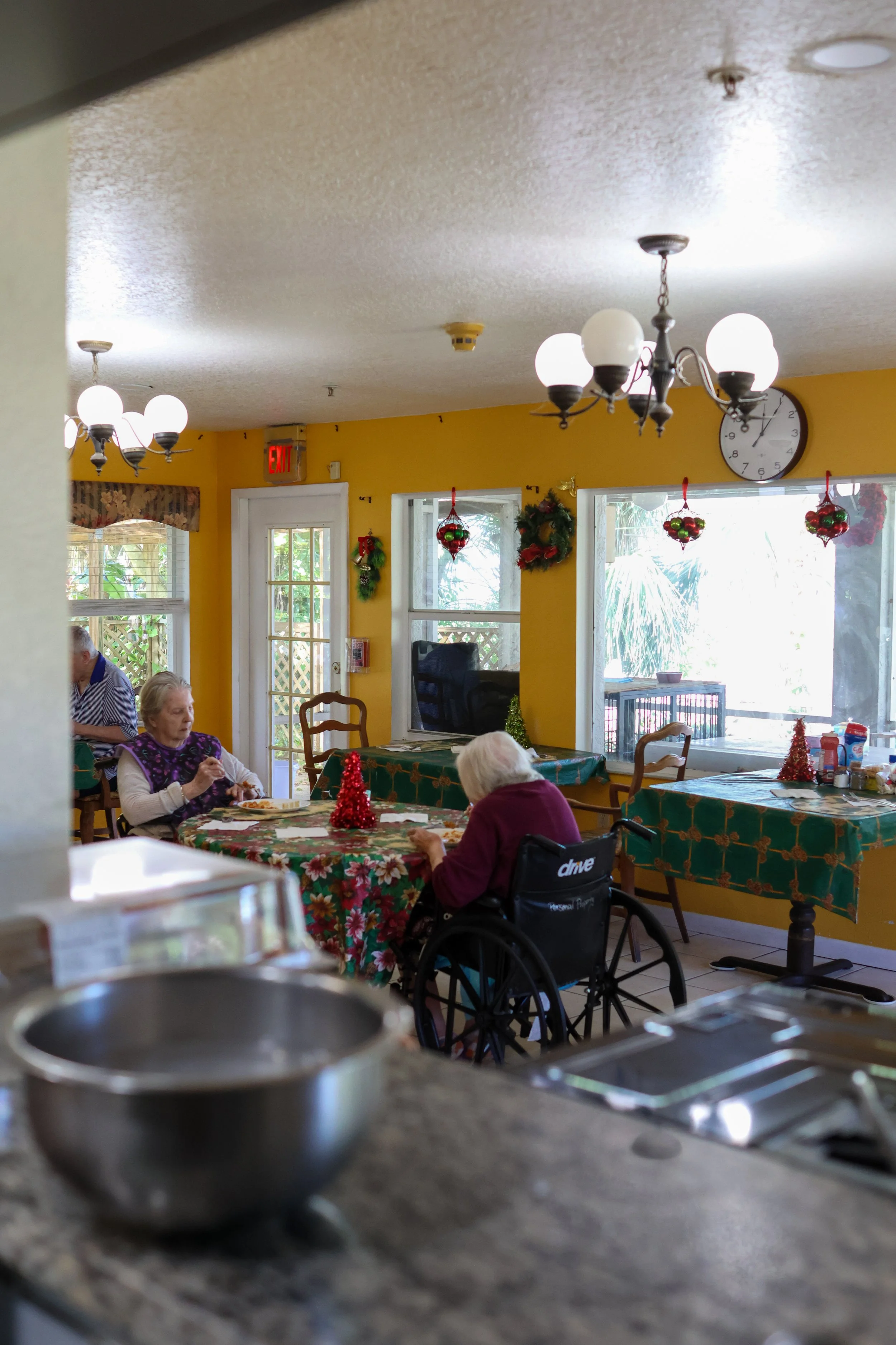 Interior of a dining area decorated for Christmas with elderly people eating at tables, some in wheelchairs, with holiday decorations hanging from the window and wall.